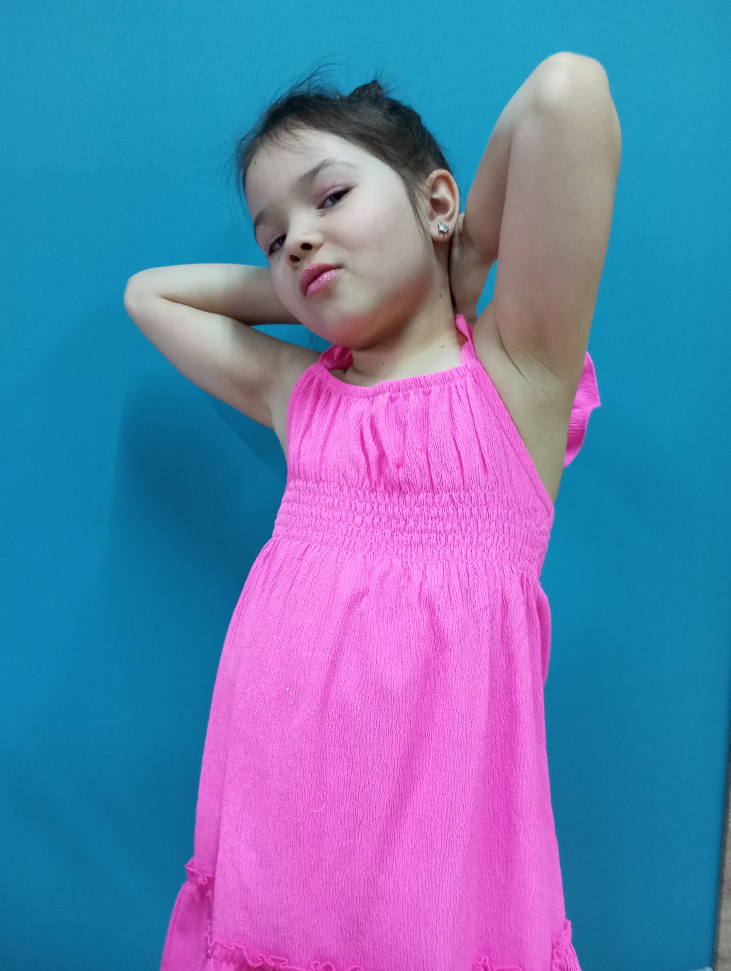 A young girl stands in front of a solid blue wall, posing with her hands behind her head. She is wearing a bright pink dress that has a gathered neckline and a crinkled texture. Her hair is pulled back in a bun, and she has a serious expression on her face. She is wearing shiny earrings. The location in Pervomaysky, Russia, is not discernible from the image.