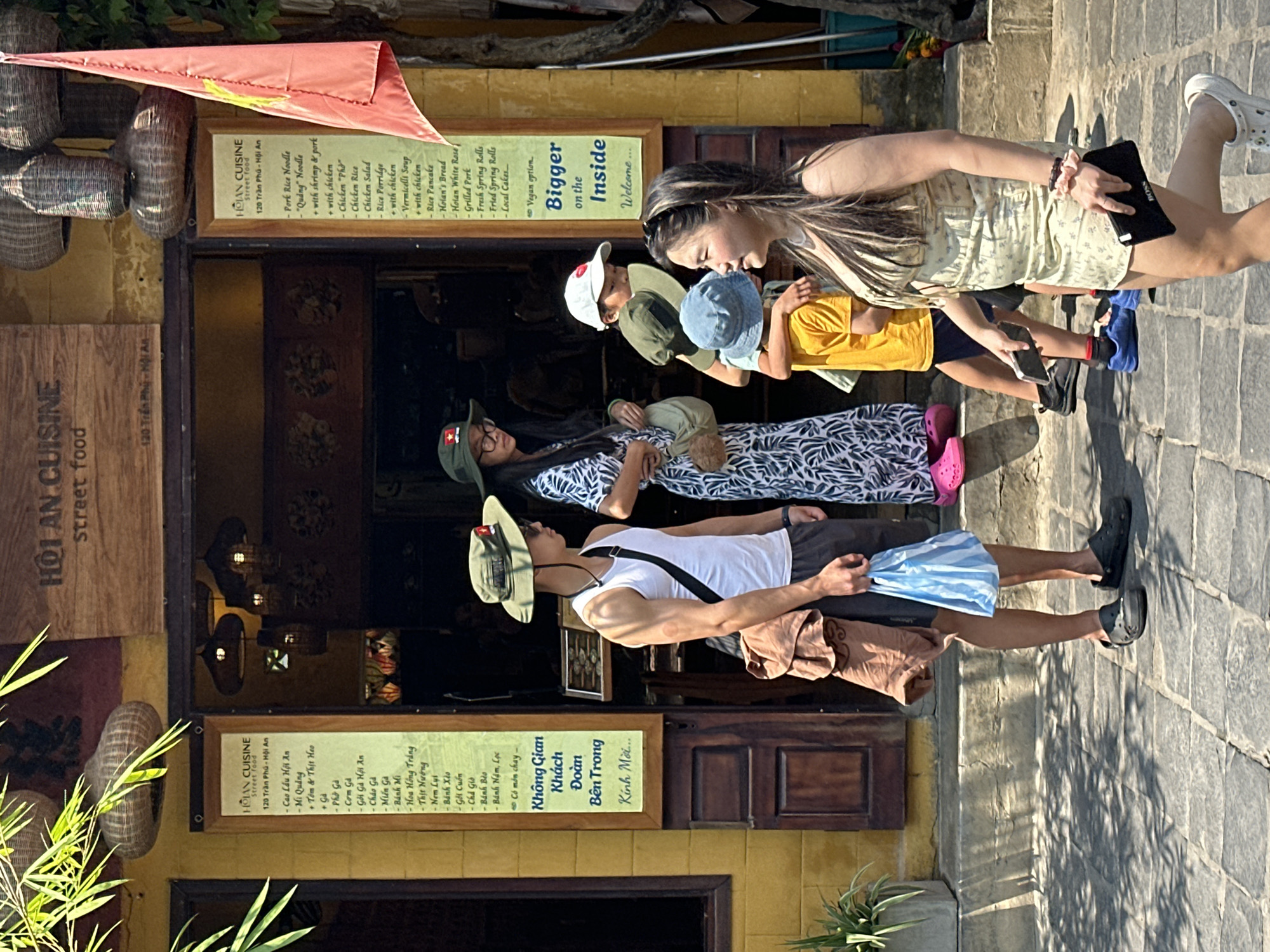 Here's a detailed description of the image:
**Location:** The image appears to be taken in Hội An Ward, Vietnam. This is indicated by the signs in front of the building, specifically noting "HỘI AN CUISINE," "120 Trần Phú - Hội An."
**Environment:** The scene is on a paved street in front of a yellow building. The building is likely a restaurant due to the menus displayed outside. There are woven items hanging near the building. The lighting suggests it is daytime.
**People:**
* A group of people is standing in front of the restaurant.
* A man with a sunhat, white tank top, brown shorts, and black footwear stands in the foreground. He is holding a blue and white striped bag.
* A woman with a sunhat with a patch on it and glasses is standing in front of another woman.
* A child with a sun hat and a yellow shirt is partially visible.
* A young woman with long hair, wearing a dress and white Crocs, is visible in the background, holding a black electronic device with the word "BOSS" on it.
**Objects:**
* Menus displaying various Vietnamese dishes.
* A Vietnamese flag hanging above one of the menus.
* Woven decorative items.
* An electronic device (likely a tablet) held by the young woman.
**Actions and Interactions:**
* The group appears to be considering the menu and deciding whether to enter the restaurant.
* The woman with long hair is looking down at her device.