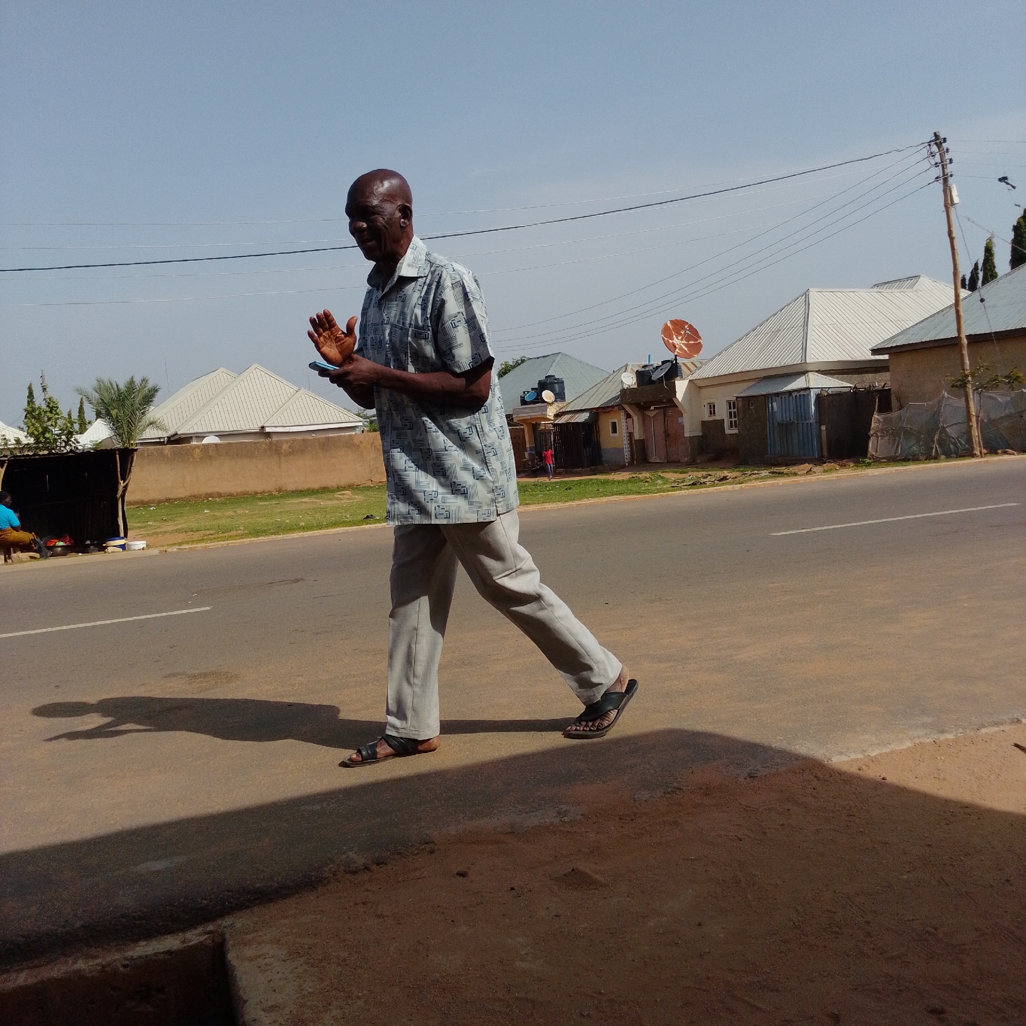 In Gombe, Nigeria, an older Black man with a shaved head is walking on a paved road. He is wearing a light blue patterned short-sleeved shirt and light grey trousers, with black sandals. His hands are raised, and he is holding a blue mobile phone in his right hand. The man is walking from left to right in the frame, and his shadow stretches out before him on the road. In the background, there are several buildings with white, corrugated metal roofs, one with a satellite dish. There are also utility poles and overhead wires visible. To the left of the road, there is a small open-air stall with a dark corrugated metal roof, and a person in a blue shirt sitting inside. The environment appears to be a semi-urban area with clear skies and bright sunlight.