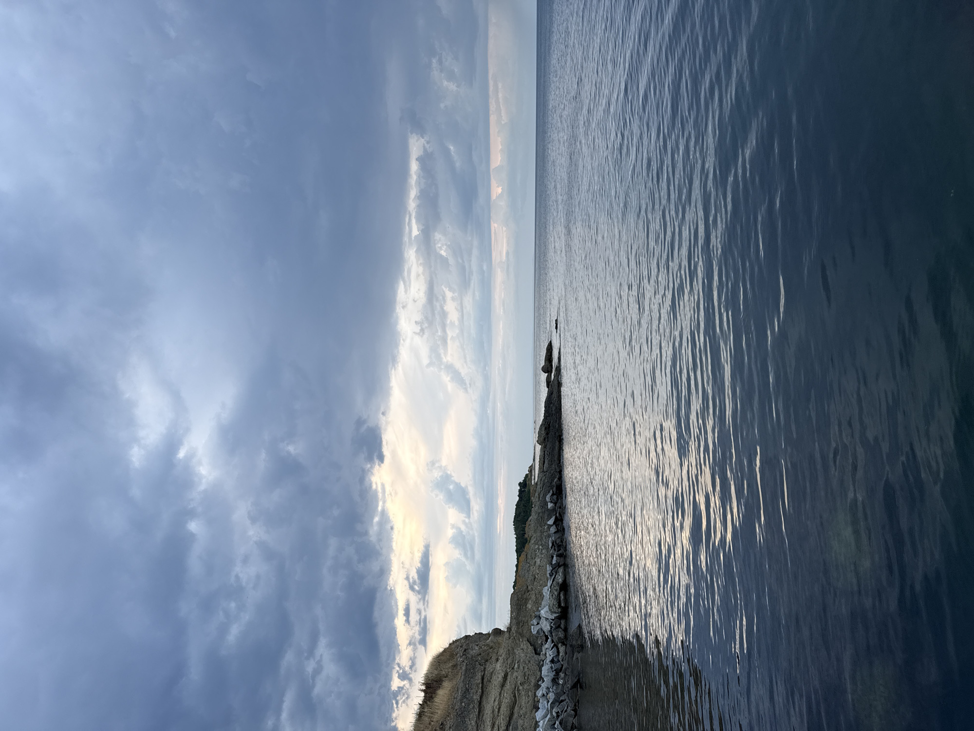 The image depicts a coastal scene near the city of Tsarevo, Bulgaria, characterized by a rocky shoreline meeting calm, dark blue water under a cloudy sky. A prominent cliff face, covered in dry, golden grass on its left side, juts out into the sea. The shoreline is a mix of natural rocks and what appears to be man-made rock barriers, extending into the water. The sea itself is smooth with gentle ripples reflecting the muted light. The sky is filled with a dramatic, overcast cloud cover, with patches of lighter sky and hints of sunset colors breaking through near the horizon. In the distance, a low, tree-covered headland is visible. There are no people or visible animals in the frame. The overall atmosphere is serene and somewhat melancholic, hinting at the approach of evening or unsettled weather.