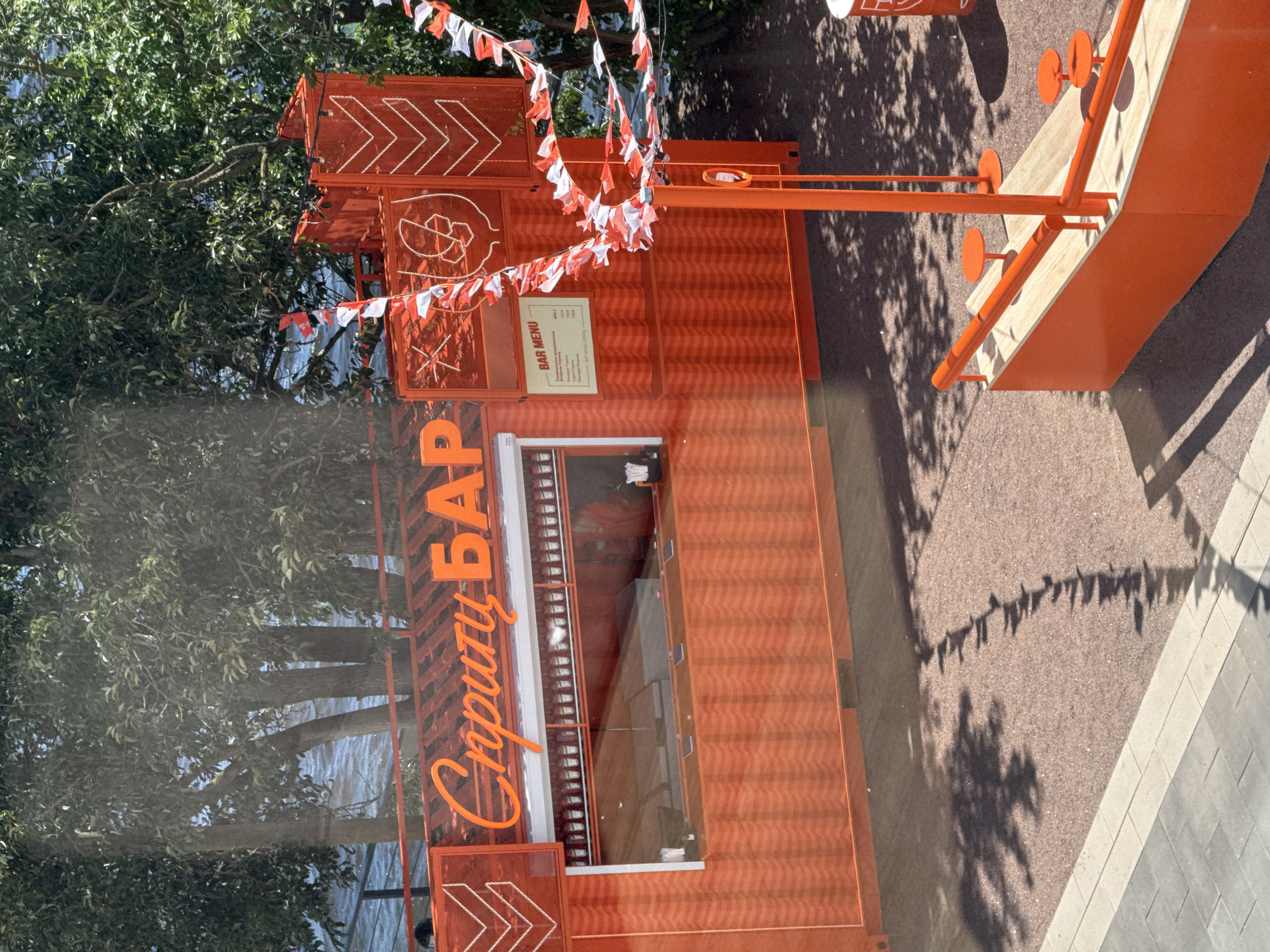 This is an outdoor, high-angle shot of a vibrant orange bar situated near a body of water, possibly a river or canal in Saint Petersburg, Russia. The bar is constructed from what appears to be shipping containers, with a large awning over the serving area and decorative fencing on the sides. Orange and white triangular flags are strung across the top of the bar structure. The bar's name, "Спиритц БАР", is displayed in orange script lettering. Inside the serving window, rows of bottles are visible. A menu is posted on the side of the bar. In front of the bar, a wooden counter area with orange railings and small orange stools is set up for customers. The ground is covered in reddish-brown gravel and there are paved pathways. Tree branches cast dappled shadows across the scene, indicating it is a sunny day. The water in the background is blue and appears to be in motion. No people are clearly visible in the image.