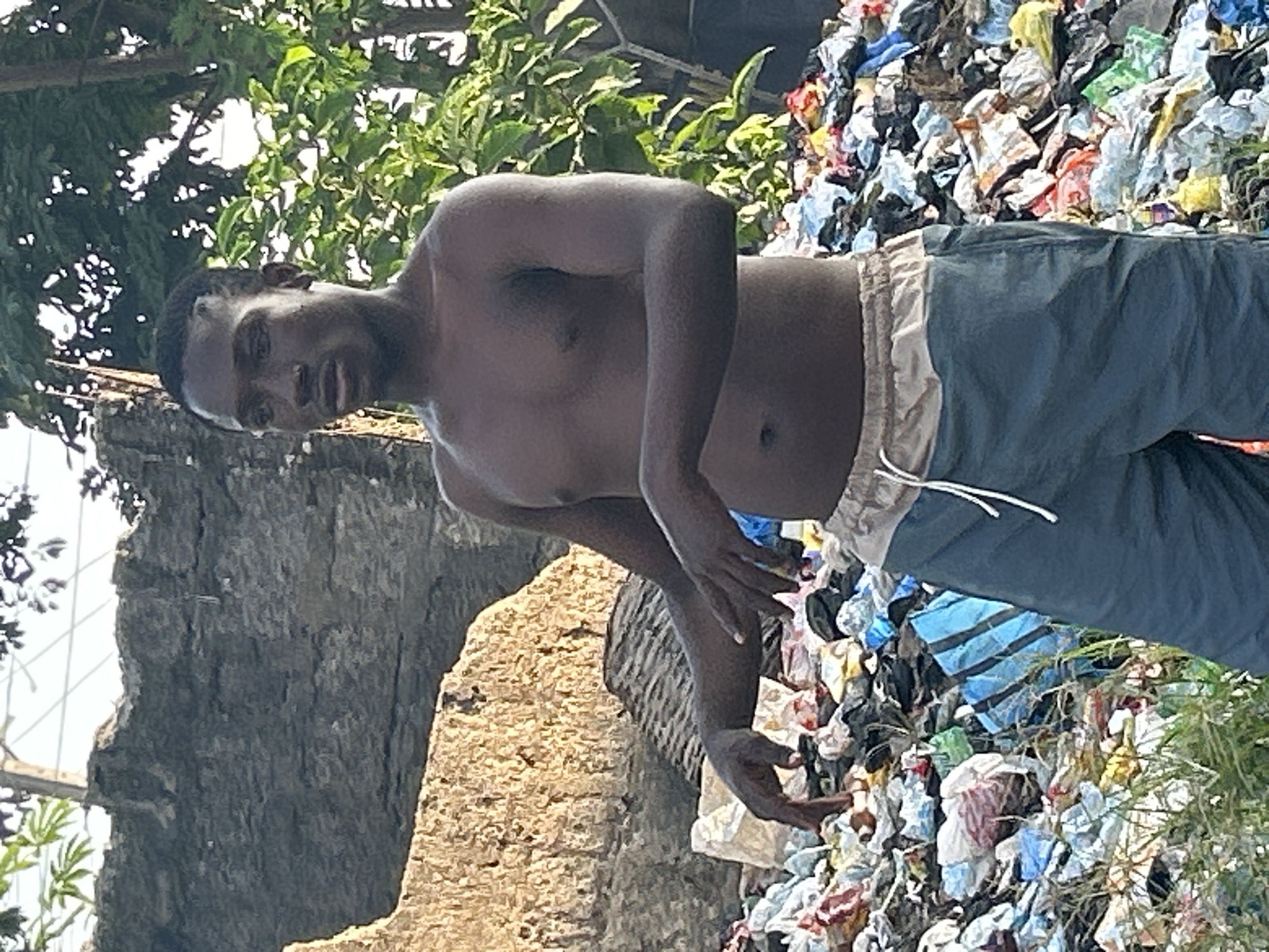 In Ihiagwa, Nigeria, a young Black man, shirtless and wearing teal shorts, stands amidst a large pile of refuse. The trash consists of various colorful plastic bags, discarded clothing, and other assorted debris, creating a chaotic and unsightly environment. Behind the man, a rough-textured, light-brown wall or rock formation is visible, partially obscured by lush green foliage. The man's posture suggests he is either observing the trash or interacting with it, with his hands extended towards the pile. The overall scene depicts an informal waste disposal site.