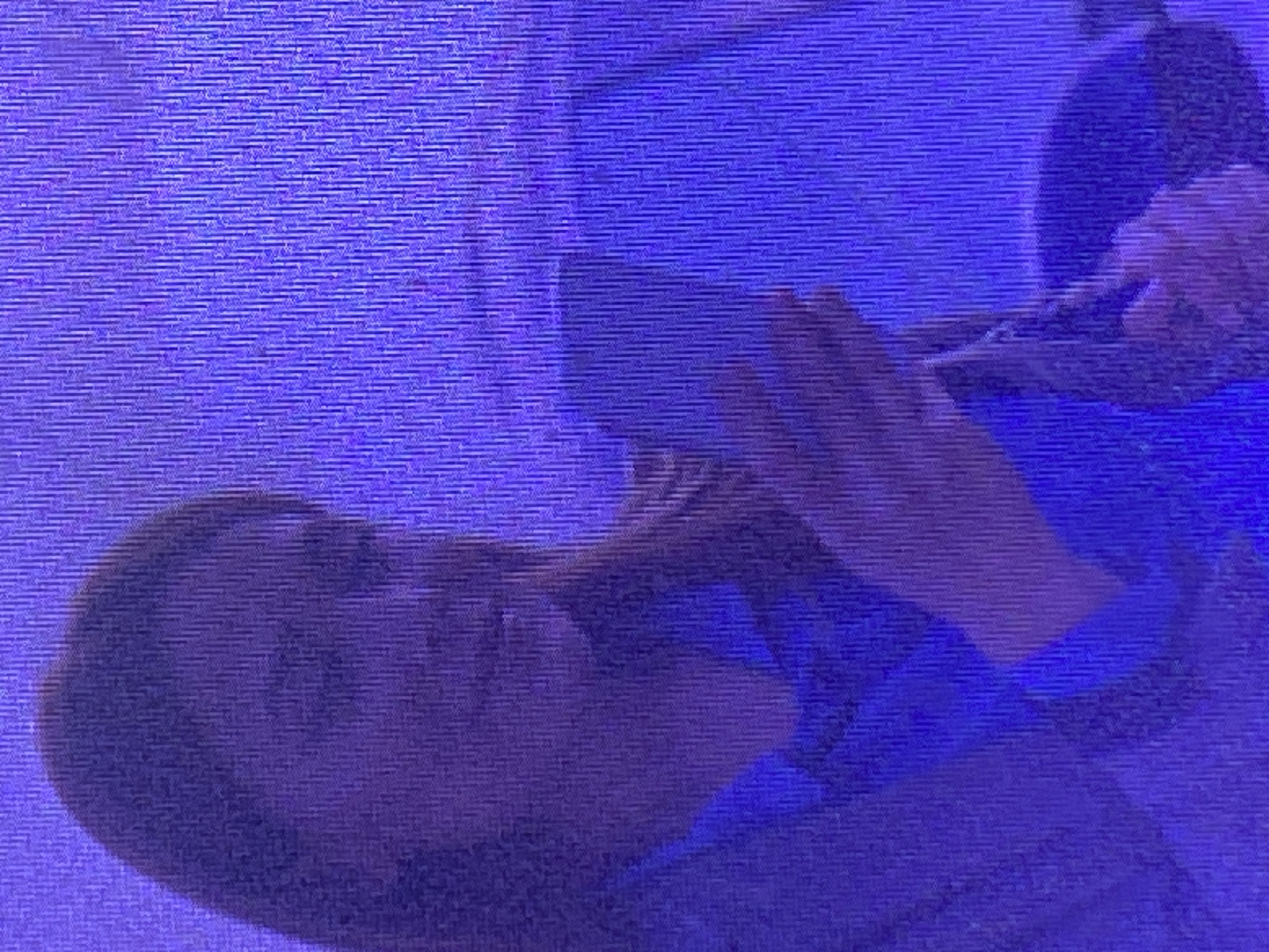 A low-resolution, purple-hued image shows a woman from the chest up, with her left side facing the camera. She is looking down at a smartphone in her hands, her face obscured by shadow. She is wearing a blue collared shirt. The background consists of a textured, purple surface, possibly a wall or fabric. There is no discernible location in Ikorodu, Nigeria, as the image lacks identifying landmarks.