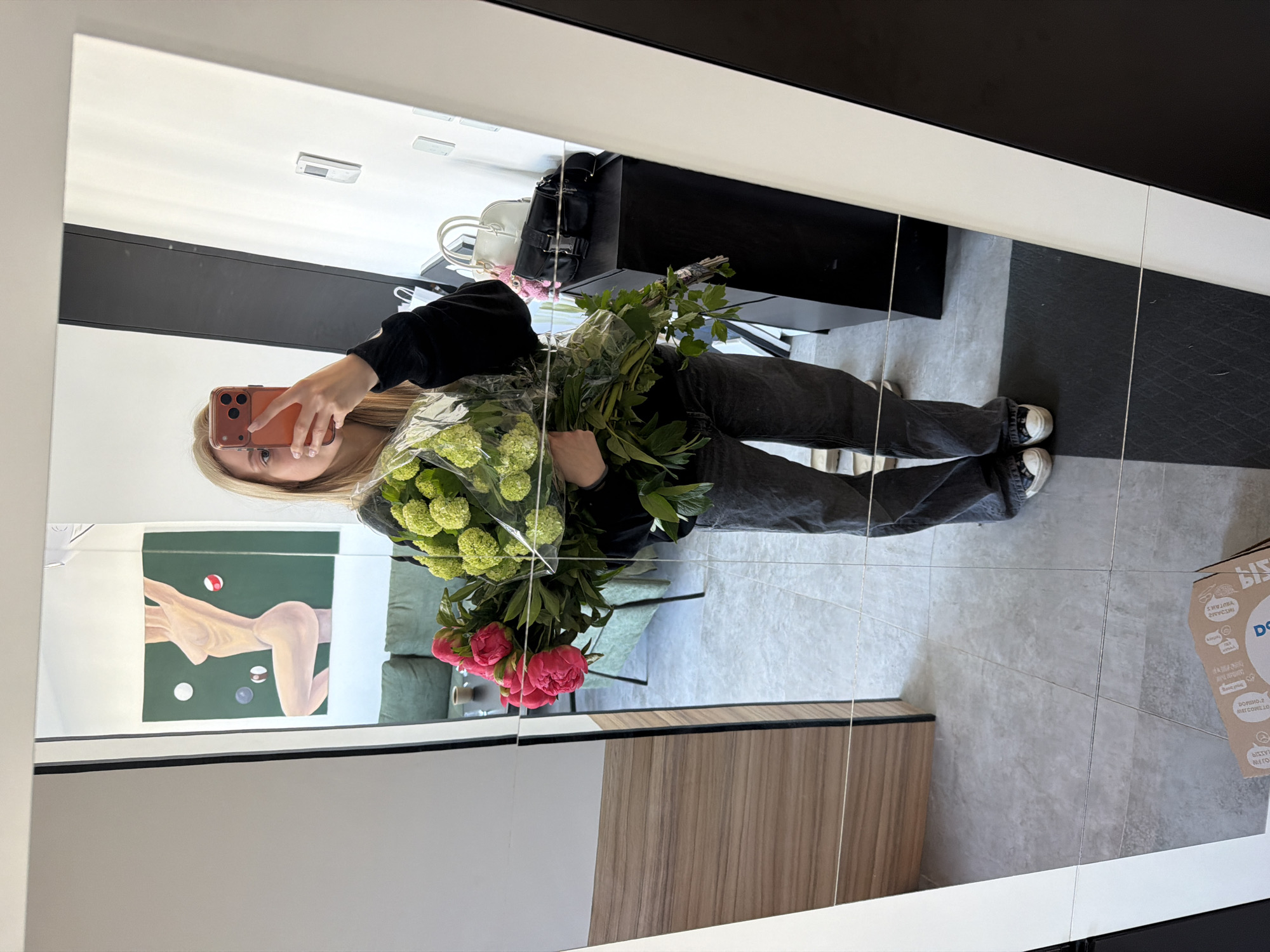 A person is visible in a mirror selfie, holding a bouquet of flowers. The individual is wearing dark pants, a black sweatshirt, and white sneakers. They are holding an orange smartphone and facing the mirror.
The reflection reveals a room with a green artwork depicting a nude figure on a green background. There's a light green couch, a black console table with two handbags, and grey tiled flooring. A cardboard box is visible near the floor.