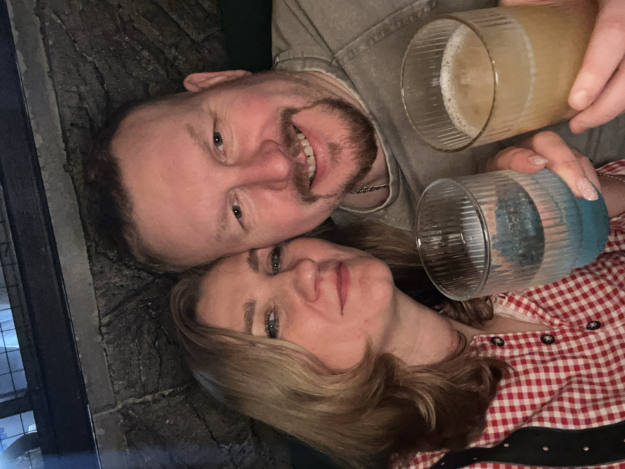 A medium shot depicts a man and a woman posing intimately with drinks in a dimly lit indoor setting.
The man, positioned on the upper left of the frame, has fair skin, short brown hair, and a full beard. He is smiling broadly, displaying his teeth. He wears a light brown, textured shirt and a thin silver chain is visible around his neck. He is holding a glass of a frothy amber-colored beverage.
The woman lies below him on the lower right, her face upturned towards the camera. She has fair skin and light brown, shoulder-length hair. She is wearing a red and white checkered shirt with black buttons. She holds a blue glass.
They are both lying on a dark surface that appears to be stone or concrete. A window and brick wall are visible on the left.
There is no explicit indication that this image was taken in Chaikovsky, Russia.