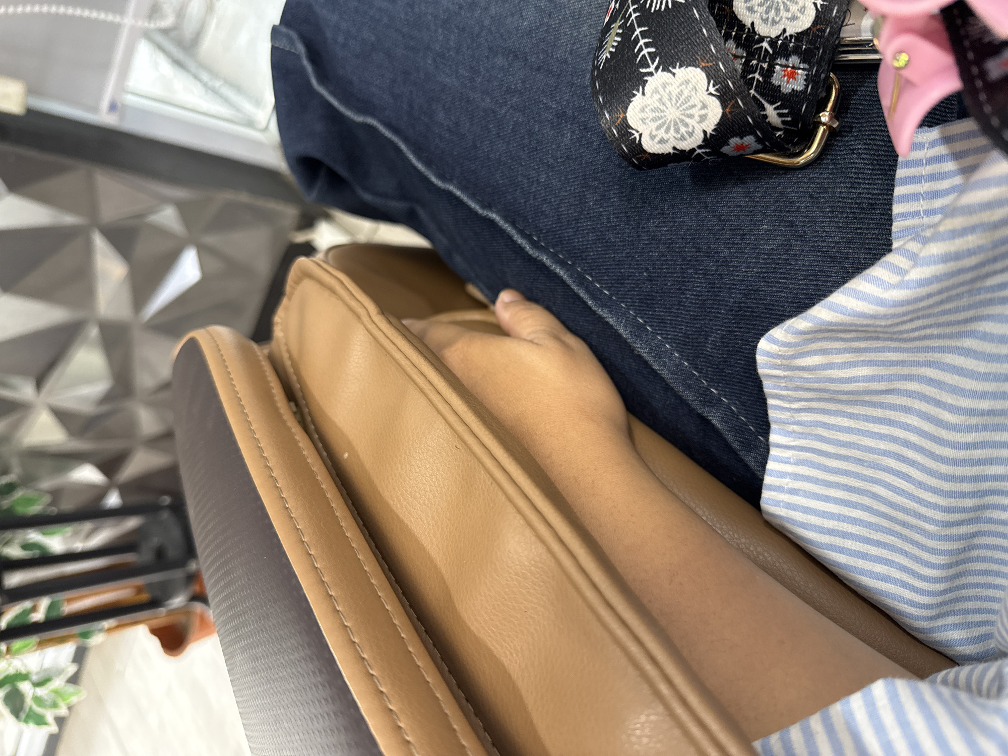 The image shows a close-up view of a person's arm and lap while seated in what appears to be a massage chair. The arm is resting on the chair's tan-colored armrest, which is made of leather. The person is wearing blue denim jeans and a blue and white striped shirt. A black fabric accessory with a floral pattern and a gold buckle is visible on the lap. The background is out of focus but suggests an indoor environment, possibly a salon or spa, with a silver textured wall and some greenery. There is no indication of the specific location within Lhokseumawe, Indonesia.