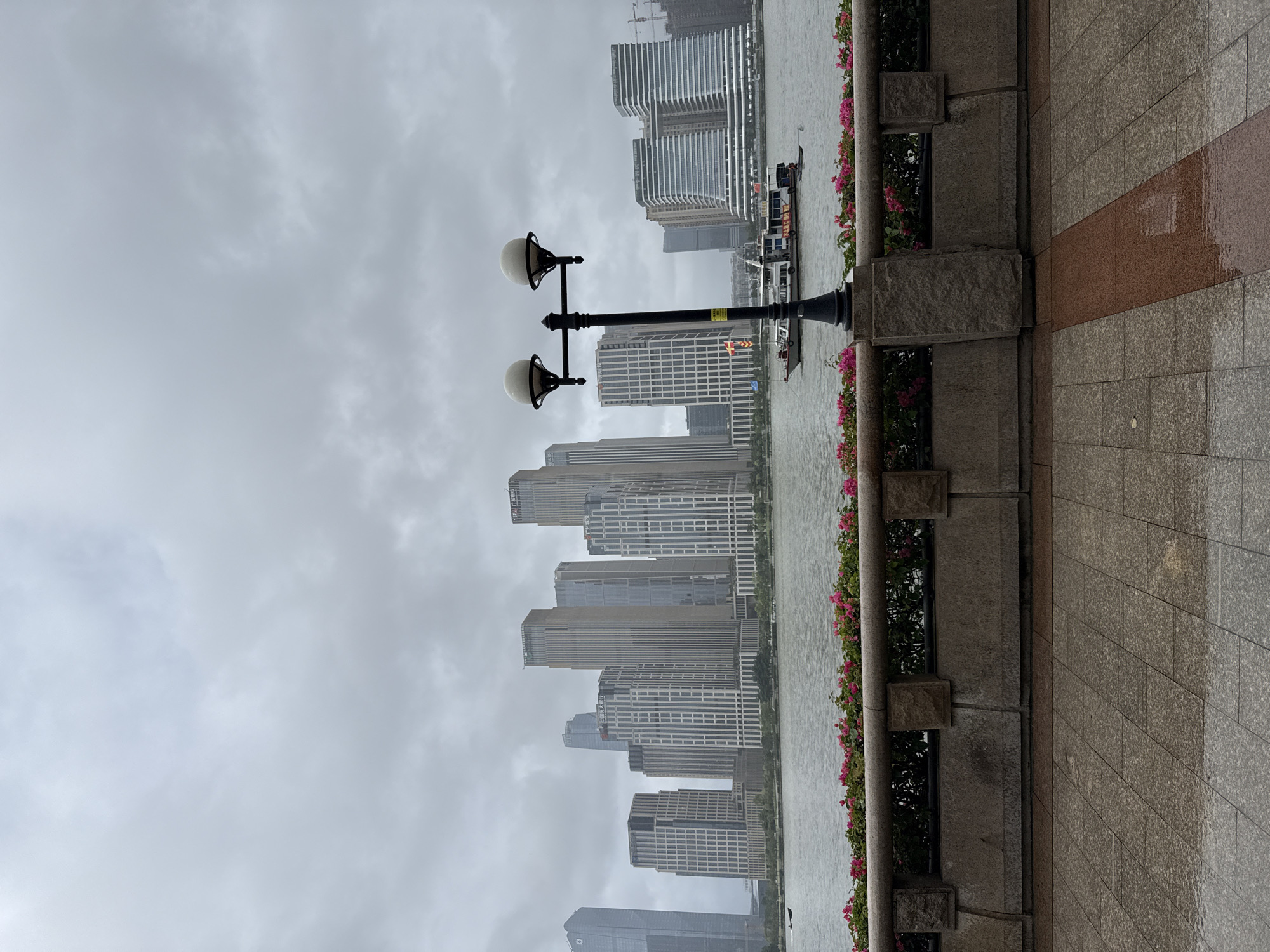 A wide shot captures a cloudy day in Haizhu District, China, with a waterfront cityscape as the focal point. The foreground is a stone pathway with a pink and brown tile pattern, appearing wet, suggesting recent rain. A low stone wall, adorned with flower boxes filled with pink flowers and dark green foliage, separates the pathway from the water. A lamppost with two spherical lights stands in the mid-ground. The river, a calm expanse of grey water, reflects the overcast sky. In the distance, a modern skyline features numerous tall buildings of varying architectural designs, mostly in shades of grey and white. A white passenger boat with red and yellow accents navigates the river, with a small dinghy behind it. Two figures are visible on the boat, and one person is in the dinghy. The sky is heavily clouded and uniformly grey.