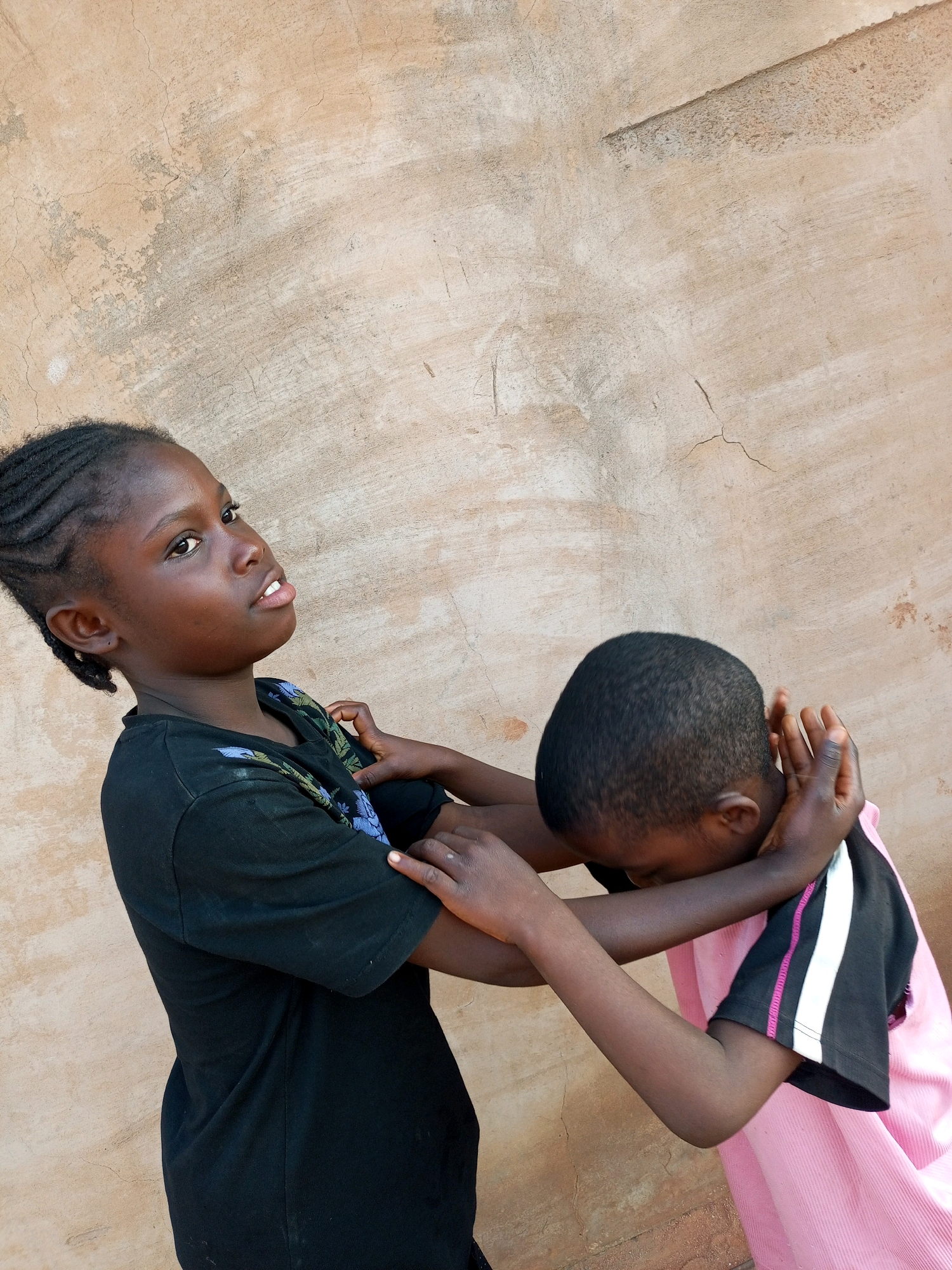 Two young people are depicted in this image. The person on the left, with braided hair and a black t-shirt, is looking to their right with a slightly raised chin. Their arms are crossed, and their hands are visible. The person on the right, wearing a pink shirt with black and white accents on the sleeves, has their head bowed and their arms are around their neck, with their hands clasped together. They are positioned to the right of the first person and are partially obscuring the wall behind them. The background is a textured, light-colored wall with visible cracks and imperfections, suggesting an outdoor setting. The image is taken from a medium distance, focusing on the two individuals and their interaction. The lighting suggests it is daytime. This scene is consistent with an outdoor environment in Tempure, Nigeria, where such informal interactions and settings are common.
