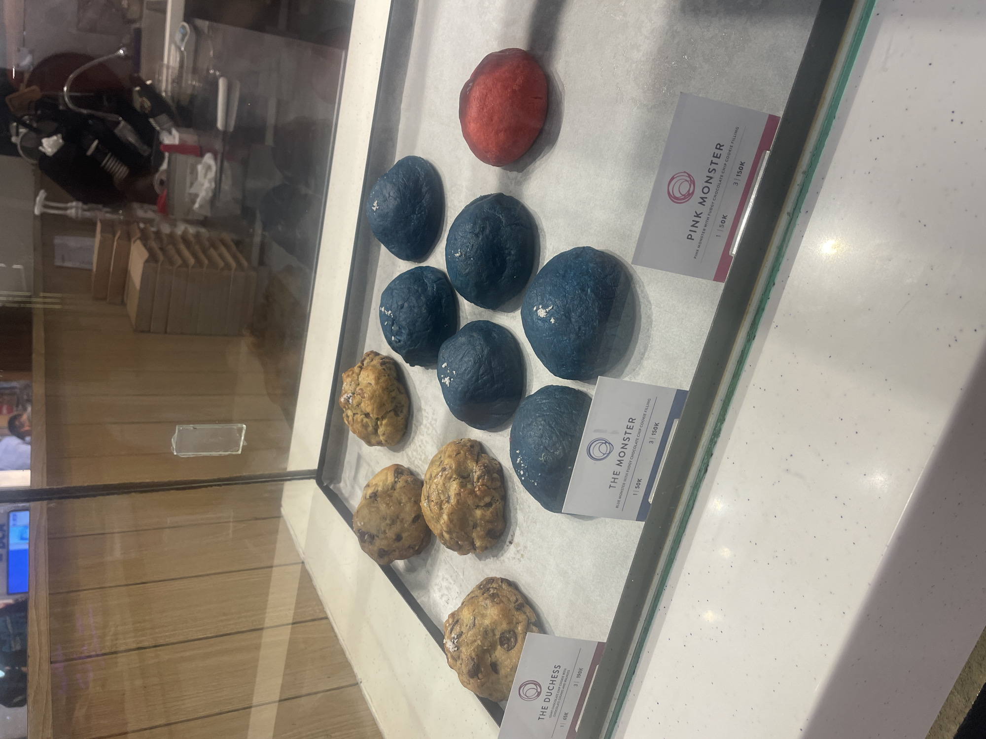 The image displays a brightly lit glass display case showcasing a variety of cookies or pastries, likely in a bakery or cafe in Denpasar, Indonesia. The items are arranged on what appears to be white parchment paper.
The top row features a single, distinct red or pink cookie, identified by an adjacent label as "PINK MONSTER." The label states, "PINK MONSTER WITH FUDGY CHOCOLATE CHIP COOKIE FILLING," priced at "1 | 50K" and "3 | 150K." Below it, several vibrant blue, somewhat irregular-shaped cookies are visible. These are labeled "THE MONSTER," described as "BLUE MONSTER WITH FUDGY CHOCOLATE CHIP COOKIE FILLING," also priced at "1 | 50K" and "3 | 150K." Towards the bottom, several classic-looking, golden-brown chocolate chip cookies are displayed. Their label reads "THE DUCHESS," described as "GIANT CHOCOLATE CHIP COOKIE WITH CHOCOLATE CHUNKS AND WALNUT," priced at "1 | 65K" and "3 | 190K." The "K" in the pricing likely refers to thousands of Indonesian Rupiah.
The glass display reflects some of the surrounding environment, which appears to be an indoor setting with wooden paneling and various blurry objects in the background, possibly kitchen equipment or shelves holding brown paper bags. The cookies themselves appear substantial and rustic, with some showing visible salt flakes or sugar crystals on their surface. The overall impression is one of a tempting selection of unique and classic baked goods available for purchase.