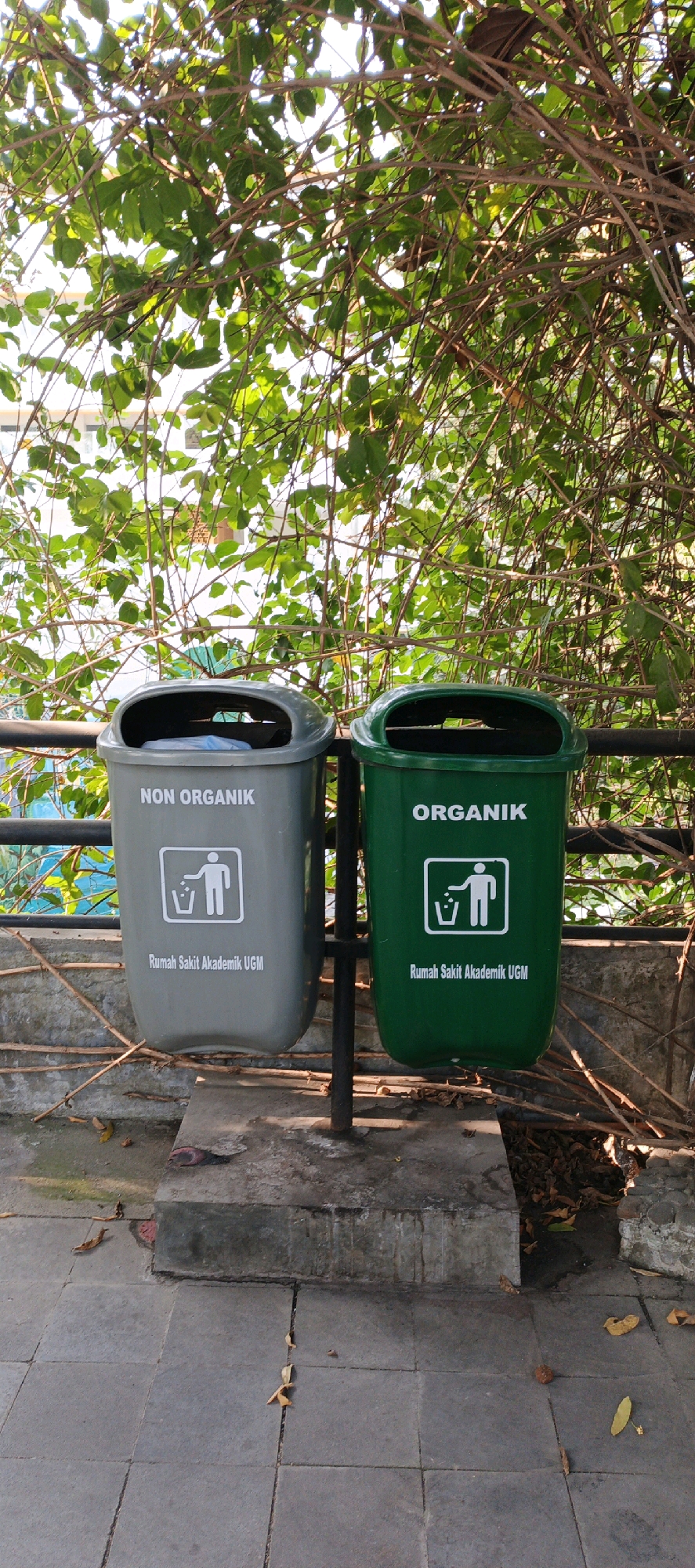 The image shows two trash cans placed on a concrete platform outdoors, with a metal railing behind them and a dense green foliage overhead. The trash cans are distinct in color and labeling. The trash can on the left is gray and labeled "NON ORGANIK" (Non-organic) in white text, with a white icon depicting a person throwing trash into a bin, and below that, "Rumah Sakit Akademik UGM" (UGM Academic Hospital). The trash can on the right is green and labeled "ORGANIK" (Organic) in white text, with an identical white icon and the same text "Rumah Sakit Akademik UGM". The ground in front of the platform is paved with gray square tiles, and scattered dry leaves are visible on the ground and the platform. The overall environment suggests an outdoor public space, possibly near a hospital given the text on the bins. The lighting indicates it is daytime, with sunlight filtering through the leaves.