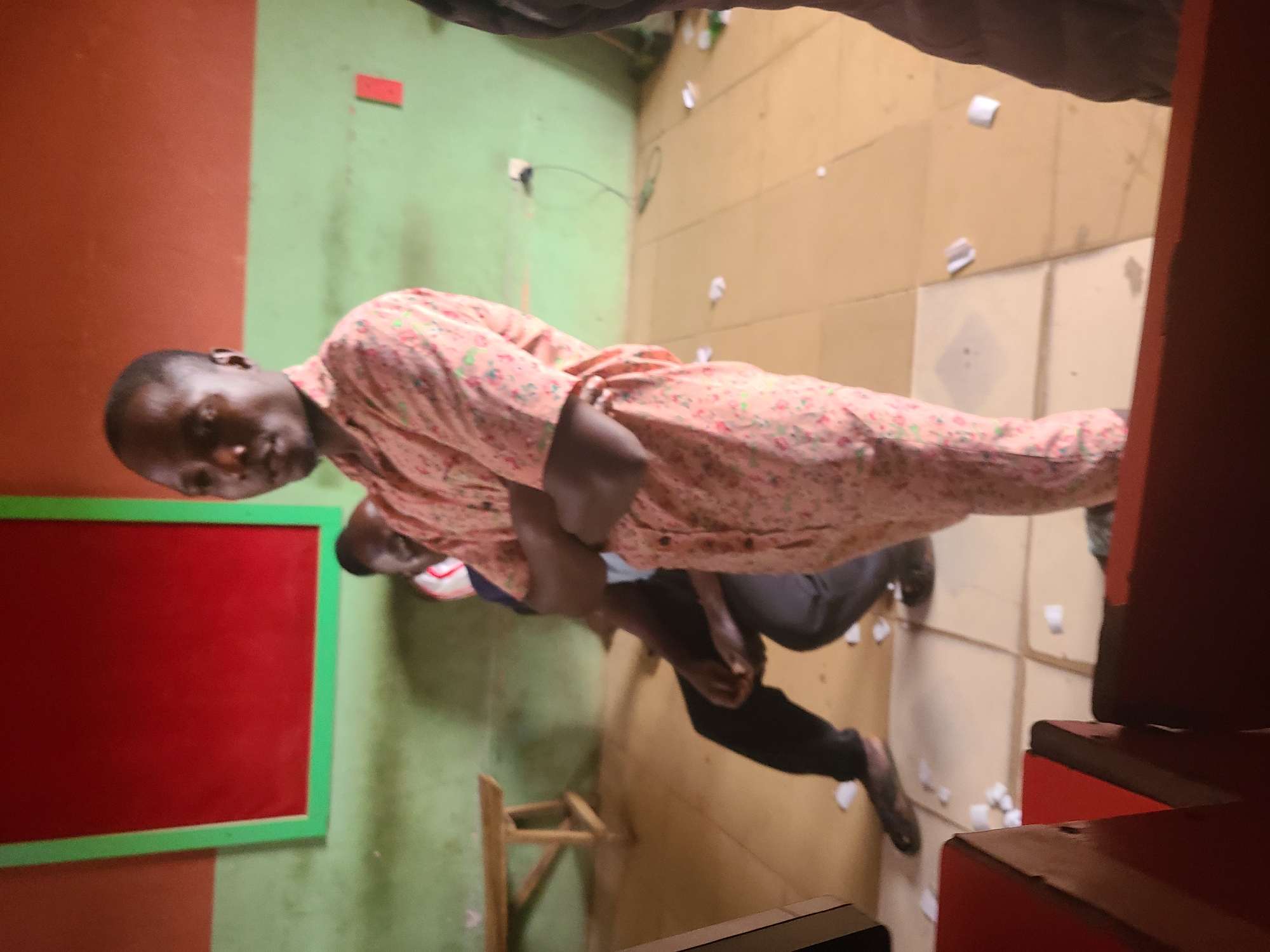 A medium shot shows a man wearing a floral-patterned pink and red shirt with black pants and dark shoes. He is positioned in a room with walls painted in two tones; one side is red, and the other is green. Behind him, another person is vaguely visible, clad in a blue shirt and darker pants. The room's floor is tiled, and scattered white paper scraps are visible. A wooden stool sits near the green wall. A red-framed chalkboard also hangs on the green and red wall. Based on the room's characteristics and lack of contextual objects, the exact location in Jos, Nigeria, is undeterminable.