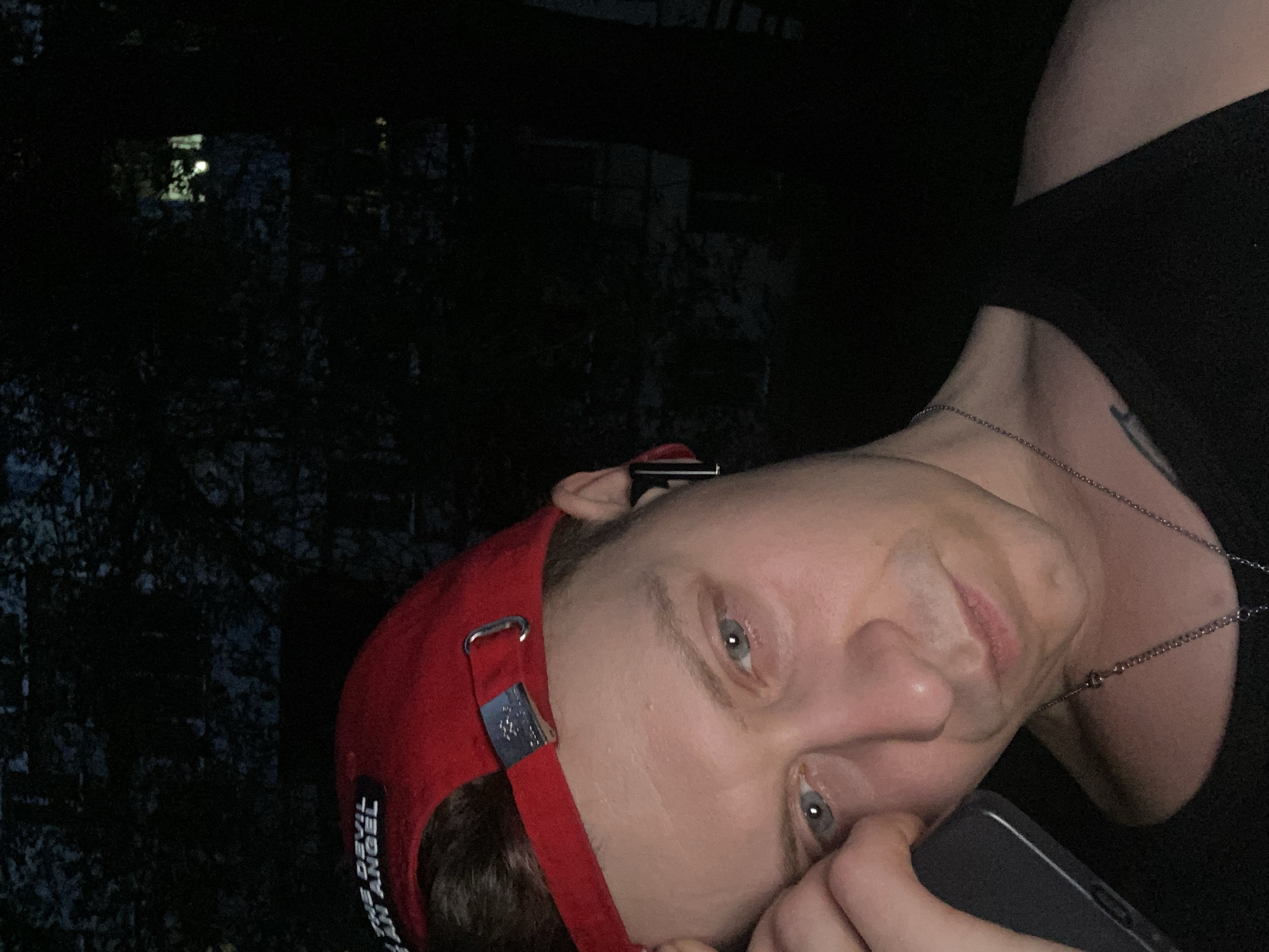 The image is a close-up, low-light selfie of a white male wearing a red baseball cap, a black tank top, and a silver chain necklace. He has light blue eyes and is resting his forehead on his hand, holding a dark-colored smartphone. An earbud is visible in his right ear. The background appears to be dark, with some indistinct foliage or trees, suggesting an outdoor setting at night. There is no discernible information in the image that would allow for the identification of the location within the city of Samara, Russia.