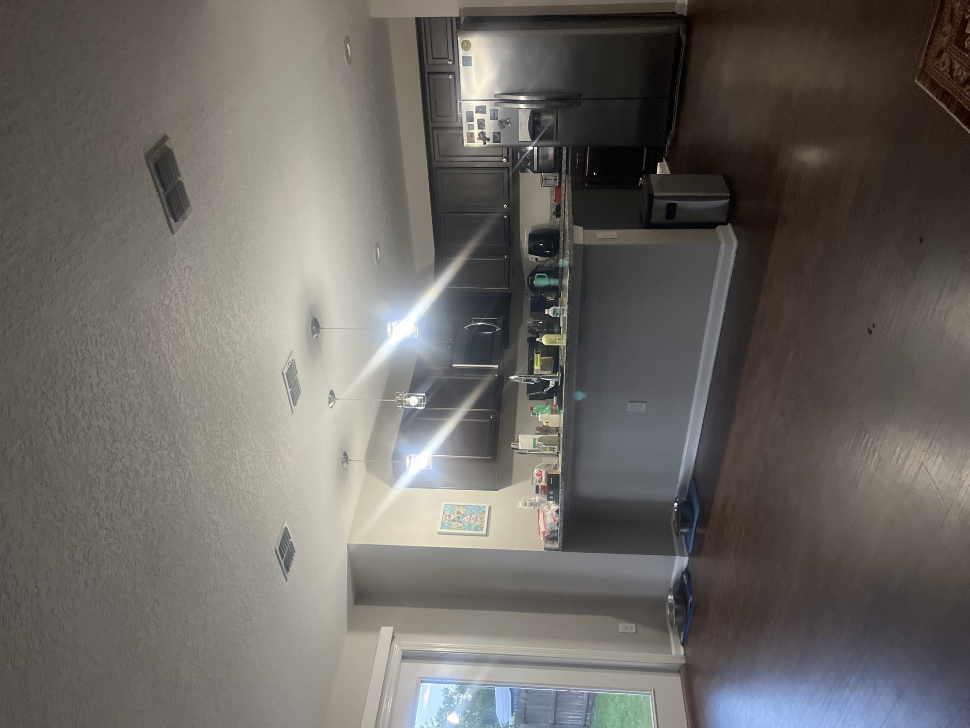 This is a wide-angle, eye-level interior shot of a modern kitchen and dining area. The flooring is dark hardwood. The left side of the image shows a glass door leading to an outdoor area with a grassy lawn and a wooden fence. Inside, near the door, are two pet food bowls on blue mats. The kitchen features dark stained cabinetry, granite countertops, and stainless steel appliances, including a refrigerator, microwave, and dishwasher. A gray island with a granite countertop separates the kitchen from the dining area. Pendant lights hang over the island. The ceiling has textured finish and includes two air vents. A silver trash can is located near the island. A framed picture hangs on the wall near the kitchen. The lighting suggests it is daytime or evening with artificial light. There are no people or animals visible in the image. The location is a residential dwelling in Jacksonville, United States of America.