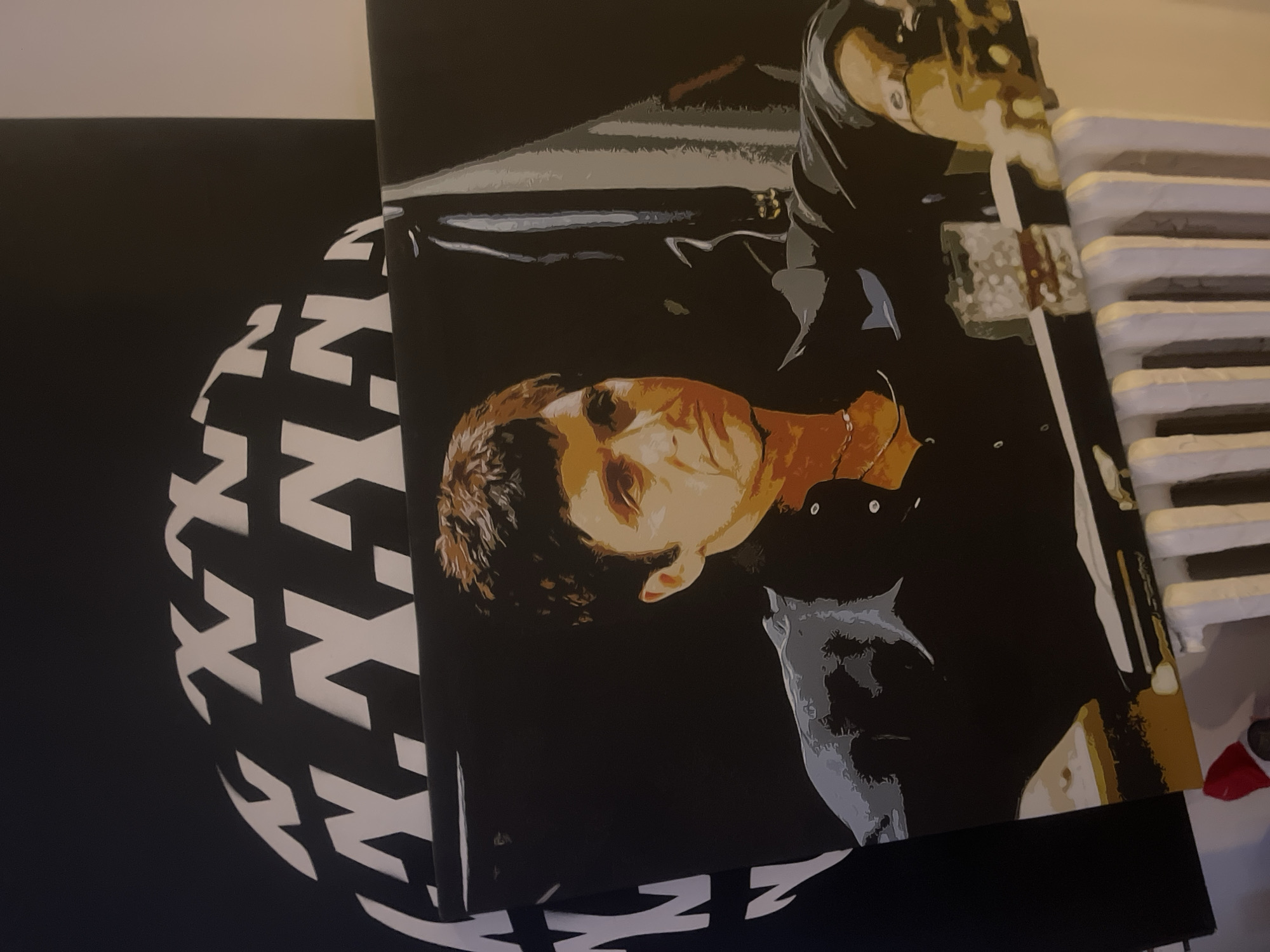 This image depicts a painting of Tony Montana, the fictional character from the film "Scarface," seated at a table with a drink. The painting is rendered in a graphic, pop-art style with bold outlines and flat colors. Tony Montana is shown wearing a black shirt and a necklace. A glass of amber liquid sits on the table in front of him. The background of the painting is black, with a suggestion of a shadowy figure or object behind him.
The painting is displayed in what appears to be an indoor setting, likely a home or gallery. Behind the painting, a large black sphere with white stylized "XX" patterns is visible. The painting itself rests on a white radiator. The lighting is somewhat dim, with a warm tone. There is no indication of a specific location in Paris, France, within the image itself.