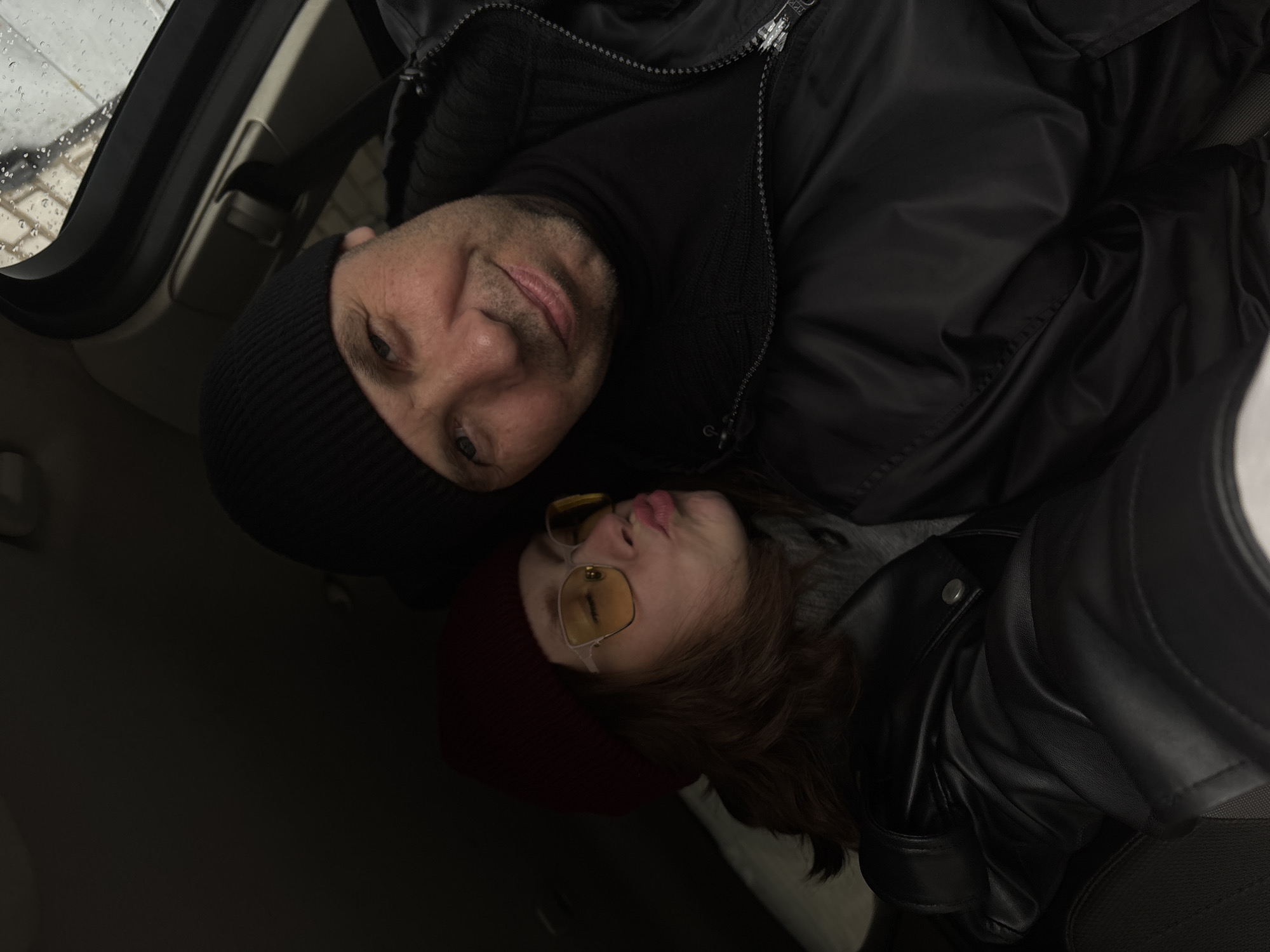 A photograph depicts two individuals, seemingly a man and a younger person, potentially a woman, posing closely together, possibly for a selfie, inside a car. The location could potentially be in the city of Chaikovsky, Russia.
The man, positioned in the upper-left portion of the frame, has short hair and is wearing a black beanie and a black turtleneck sweater. He appears to be smiling faintly.
The younger person, located in the lower-right portion of the frame, is wearing a red beanie, yellow-tinted sunglasses, and a black leather jacket. Their lips are pursed in a possible kissing gesture.
The car's interior is mostly obscured, with the windshield visible in the upper-left corner displaying raindrops. The remainder of the frame is dominated by the dark clothing of the subjects and the darkened car interior.