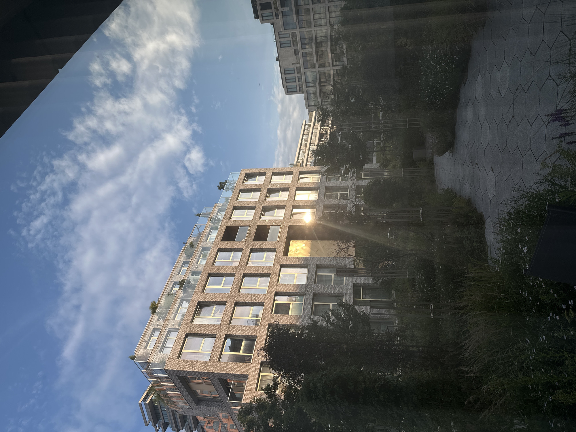 The image depicts a modern residential building in Amsterdam, Netherlands, bathed in the warm glow of the setting sun. The building, constructed with a combination of light-colored brick and glass, features multiple stories with balconies and large windows. The sun is low in the sky, casting a bright, golden reflection on a section of the building's facade and creating lens flare. In the foreground, a pathway made of hexagonal concrete pavers winds through a lush garden with various types of greenery, including tall grasses and flowering plants. Other buildings, also modern in design with glass balconies, are visible in the background, suggesting an urban setting with contemporary architecture. The sky is blue with scattered white clouds. There are no people visible in the scene.