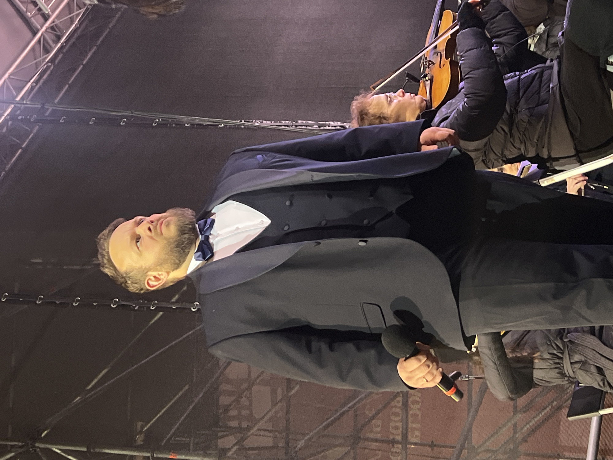 A man in a tuxedo with a blue bow tie is holding a microphone and speaking. There is a violinist behind him playing a stringed instrument. The location is Saint Petersburg, Russia.