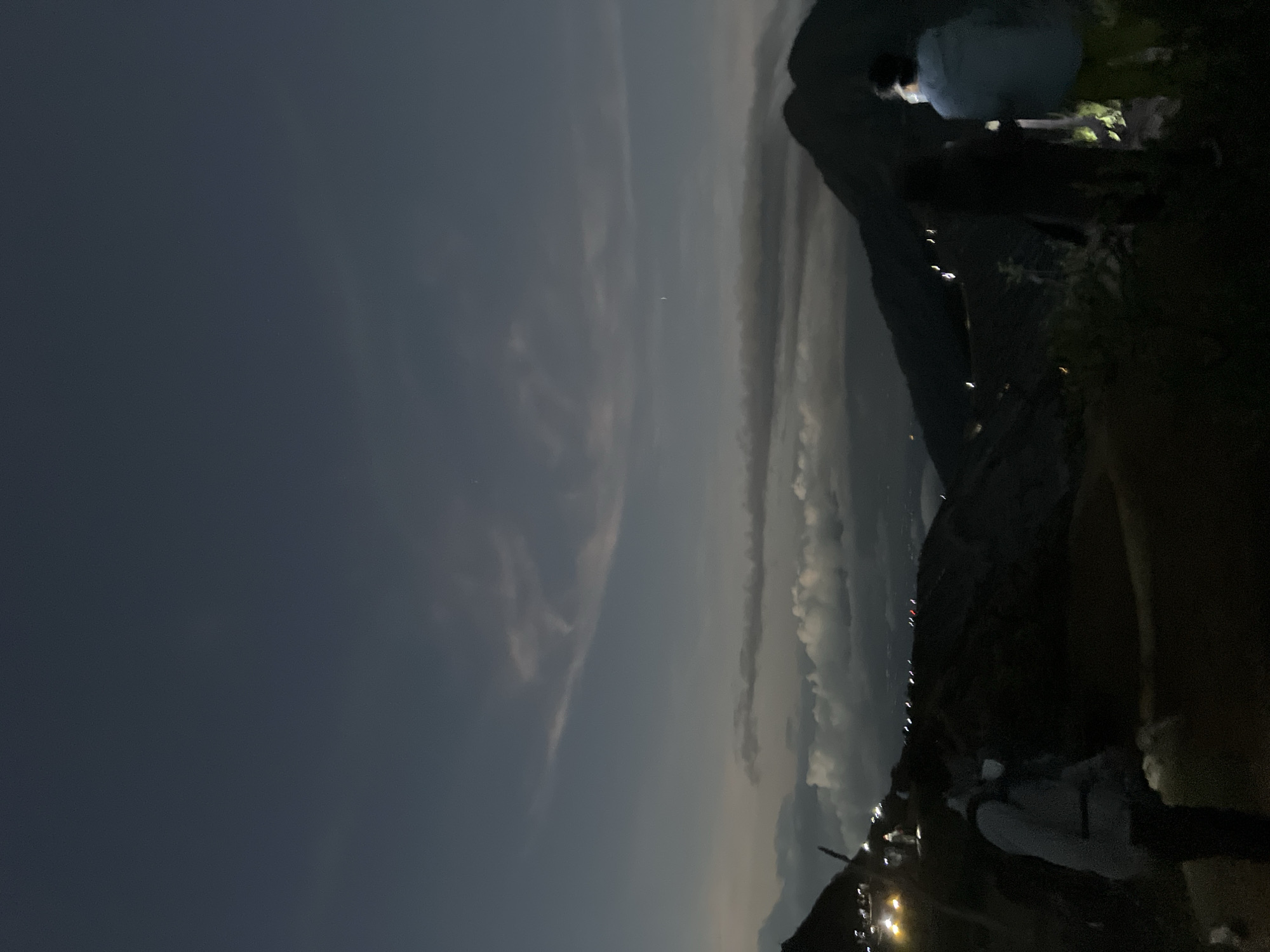 The image depicts a landscape at night in an unidentified city in Indonesia. The sky is mostly dark blue, with patches of clouds illuminated by the moon. The city lights glow faintly below a dark mountain range. A group of people are gathered near the peak of one mountain, one with a tent. The scene appears to be captured from a high vantage point, possibly another mountain or hill overlooking the city.