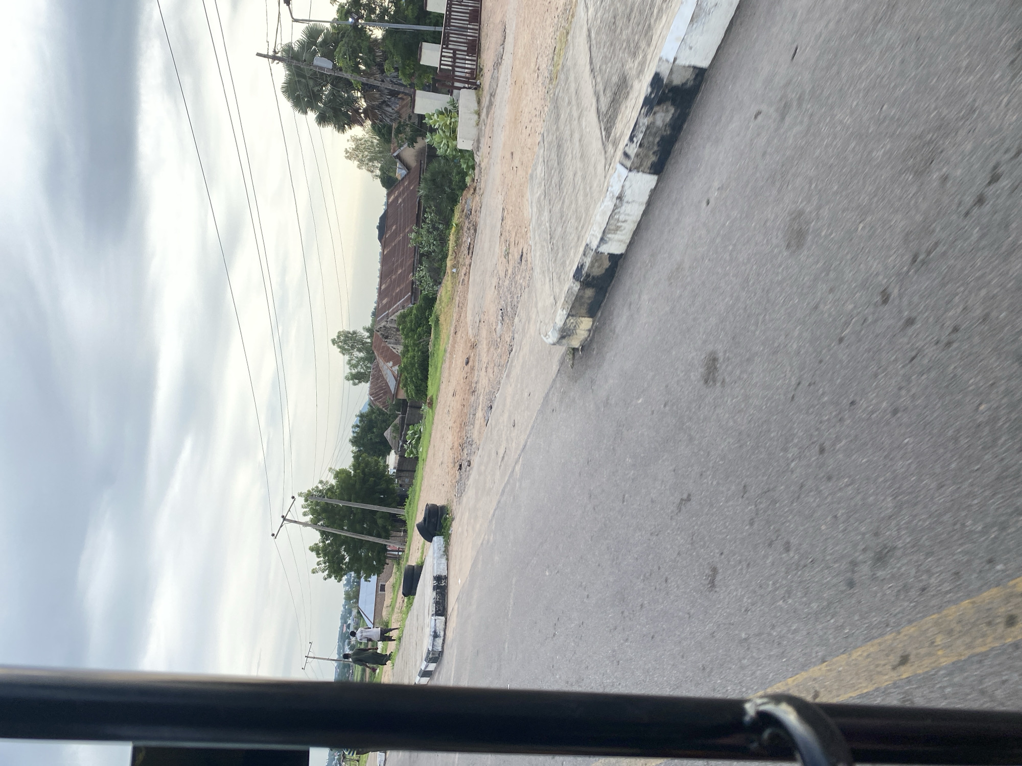 The image depicts a street scene in Jalingo, Nigeria. In the foreground, a dark pole from a vehicle is visible on the left, obscuring part of the asphalt road which has a yellow line marking. Two individuals are walking away from the camera on the right side of the road. Behind them, a raised concrete structure with black and white stripes runs along the road, with several old tires placed near it. Further back, a collection of buildings with rusty corrugated metal roofs are visible, interspersed with trees and vegetation. Power lines are strung across the sky, supported by poles. The sky is overcast with cloudy conditions. The environment suggests a residential or semi-rural area within the city.