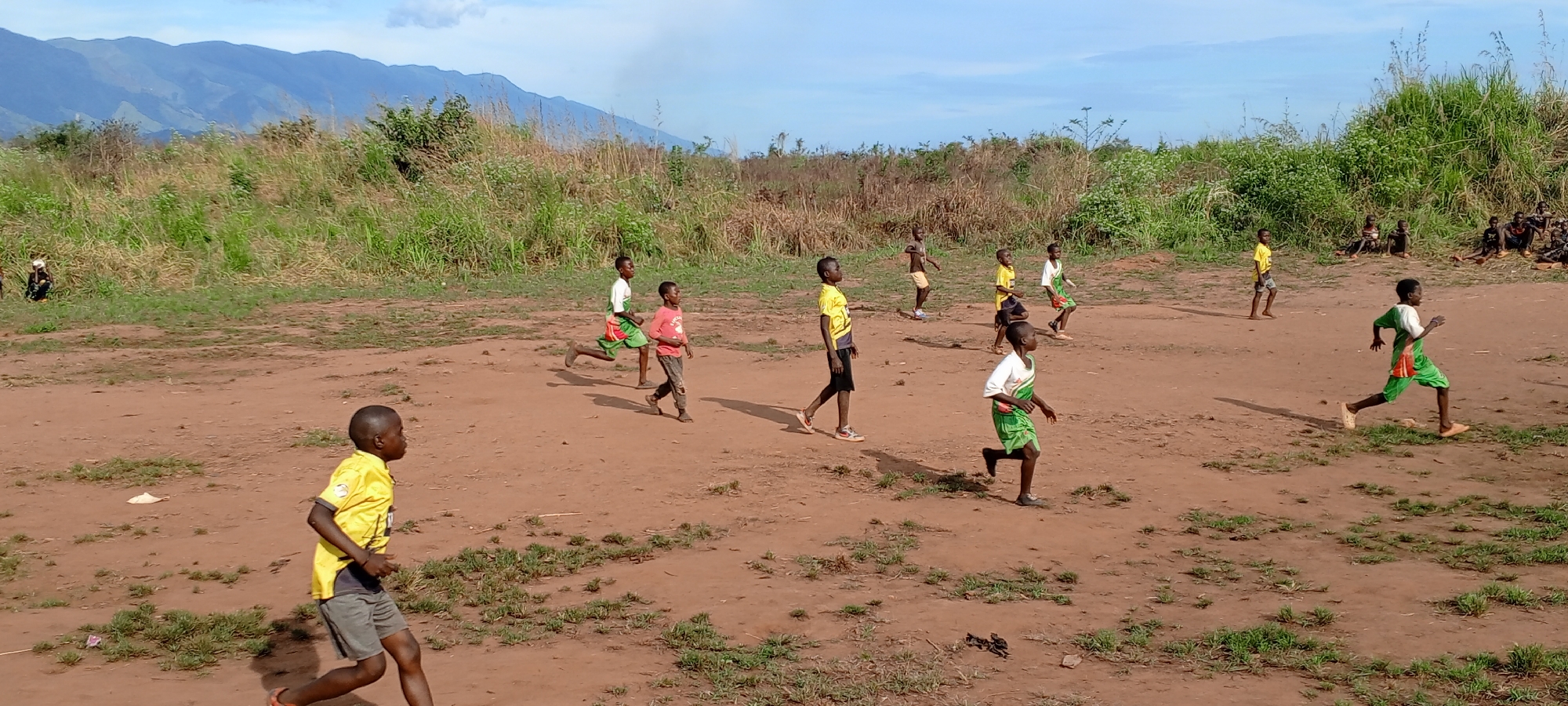 In Ngalula, Democratic Republic of the Congo, a group of children are playing on a dirt field. They wear various shirts and shorts. In the background are grassy areas and distant mountains. Some children are sitting along the edge of the field, watching the others play.