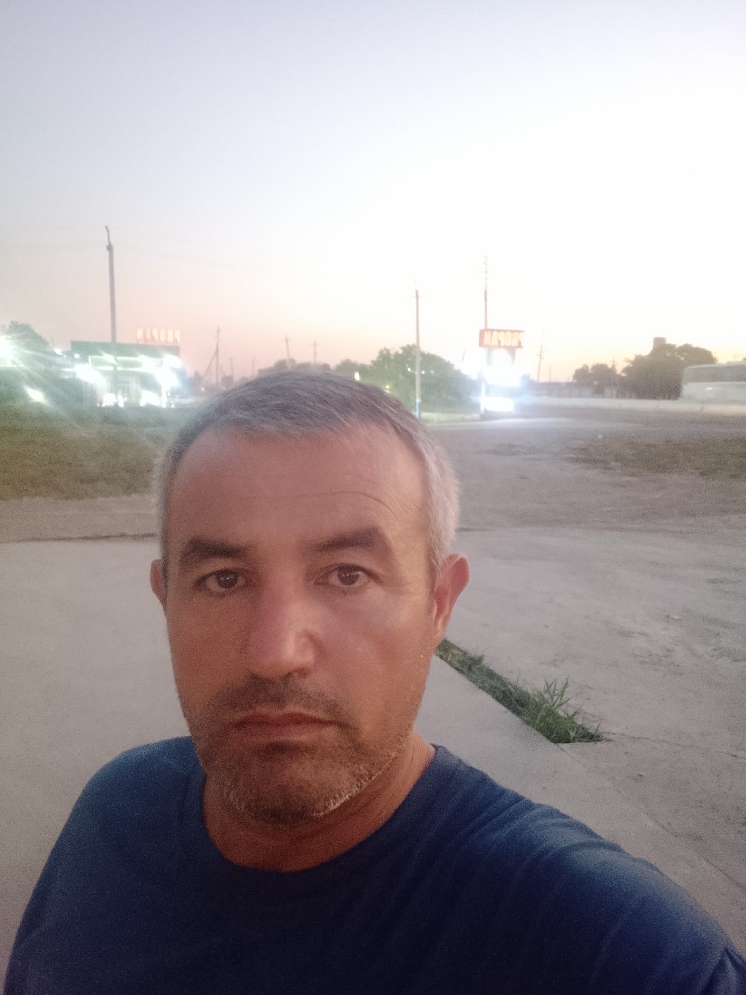 A medium-shot selfie taken from the chest up shows a middle-aged man with graying hair and stubble. He is looking directly at the camera with a neutral expression. He is wearing a dark blue t-shirt. In the background, there is a blurred gas station with illuminated signs. The sky is a pale gradient of pink and gray, suggesting it is either dawn or dusk. The overall setting appears to be an outdoor, possibly rural or suburban, area. Based on the presence of the gas station and the environment, the location is identified as Sabir Rakhimov, Uzbekistan.