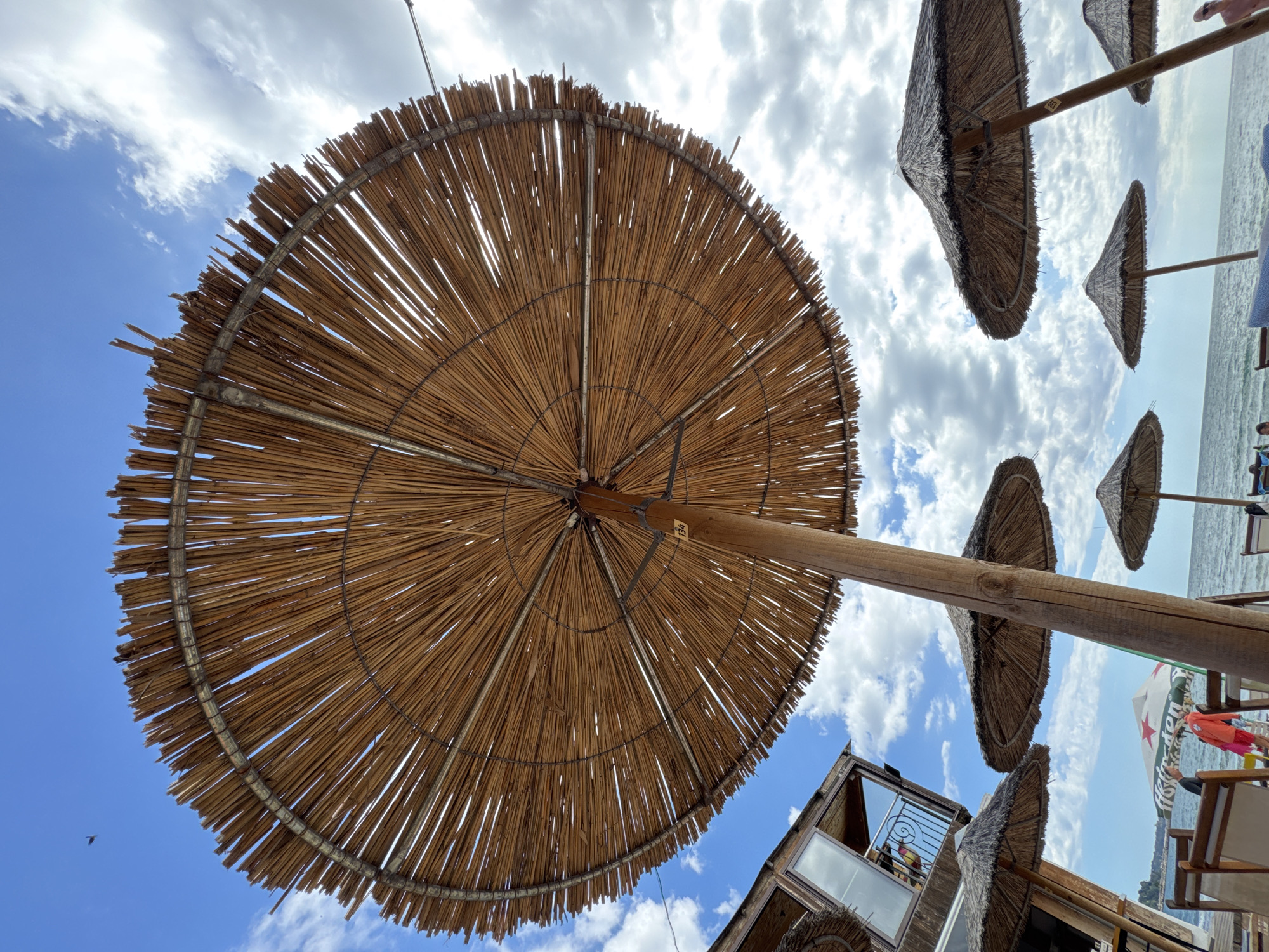 The image captures a low-angle view of a beach scene, likely in the city of Varna, Bulgaria, dominated by a large, rustic thatched umbrella in the foreground. The umbrella's natural straw material and wooden pole are clearly visible, creating a textured canopy against a bright blue sky dotted with scattered white clouds.
To the left, a portion of a building, possibly a beach bar or restaurant, is visible. It features a modern, angular design with large glass windows and a balcony with a metal railing. Several other thatched umbrellas of various sizes are scattered across the beach, some in use and others empty. In the background, the sea stretches out towards the horizon, with small waves visible.
A few people are present on the beach, some appearing to be seated on chairs or lounging on towels, while others are standing. One person is wearing a bright orange shirt, and another is wearing a pink garment. Beach chairs are positioned on a wooden deck or walkway leading towards the water. A partially visible beach umbrella with a prominent red star logo and text, possibly from a beverage company, is also in the scene. The overall atmosphere is one of a relaxed day at the seaside.