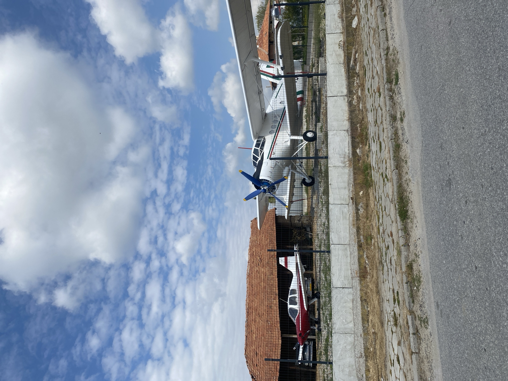 The image shows two planes and a partial view of a road. The first plane is a white, single-engine biplane with a blue propeller. The second plane is a red and white single-engine aircraft with a propeller. Both planes are displayed behind a fence. There is a red-tiled roof between them, suggesting a building. The planes appear to be located in Kapatovo, Bulgaria. The sky is visible in the background, filled with white clouds.