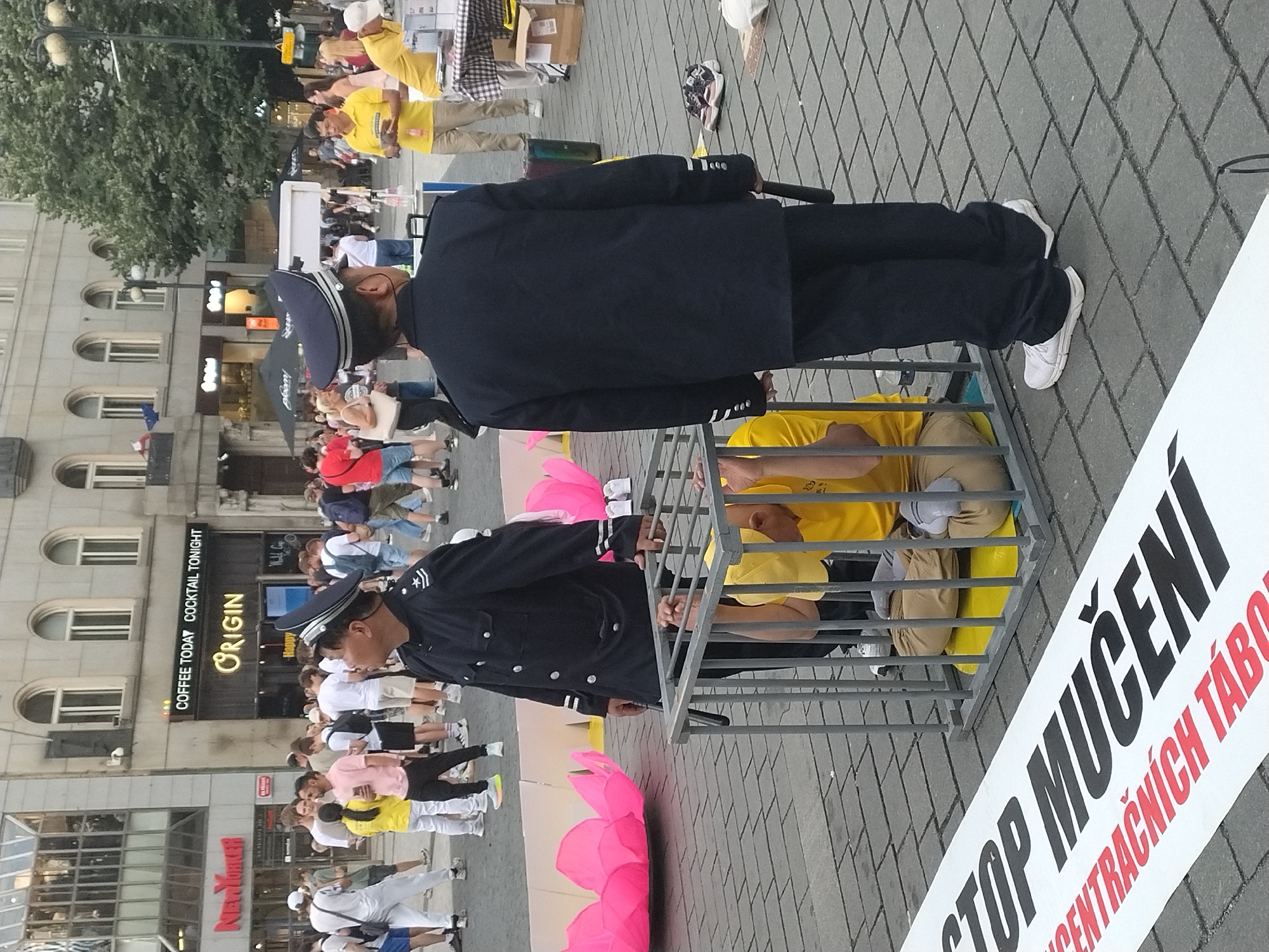 In Prague, Czechia, a protest is taking place on a cobblestone plaza. Two police officers in uniform stand guard. One officer, in the foreground and facing away from the camera, wears a dark blue uniform with a cap and holds a baton. He stands near a metal cage containing a person in a yellow shirt and tan pants, who is seated inside. Another officer, to the left of the cage and facing left, also wears a dark blue uniform and cap. A large banner in the foreground reads "STOP MUČENÍ" in bold black letters and "CENTRAČNÍCH TABO" in smaller red letters below. In the background, a crowd of people are visible, some holding banners and others observing the scene. Buildings with shops and cafes line the plaza. Large pink lotus flower decorations are also present.