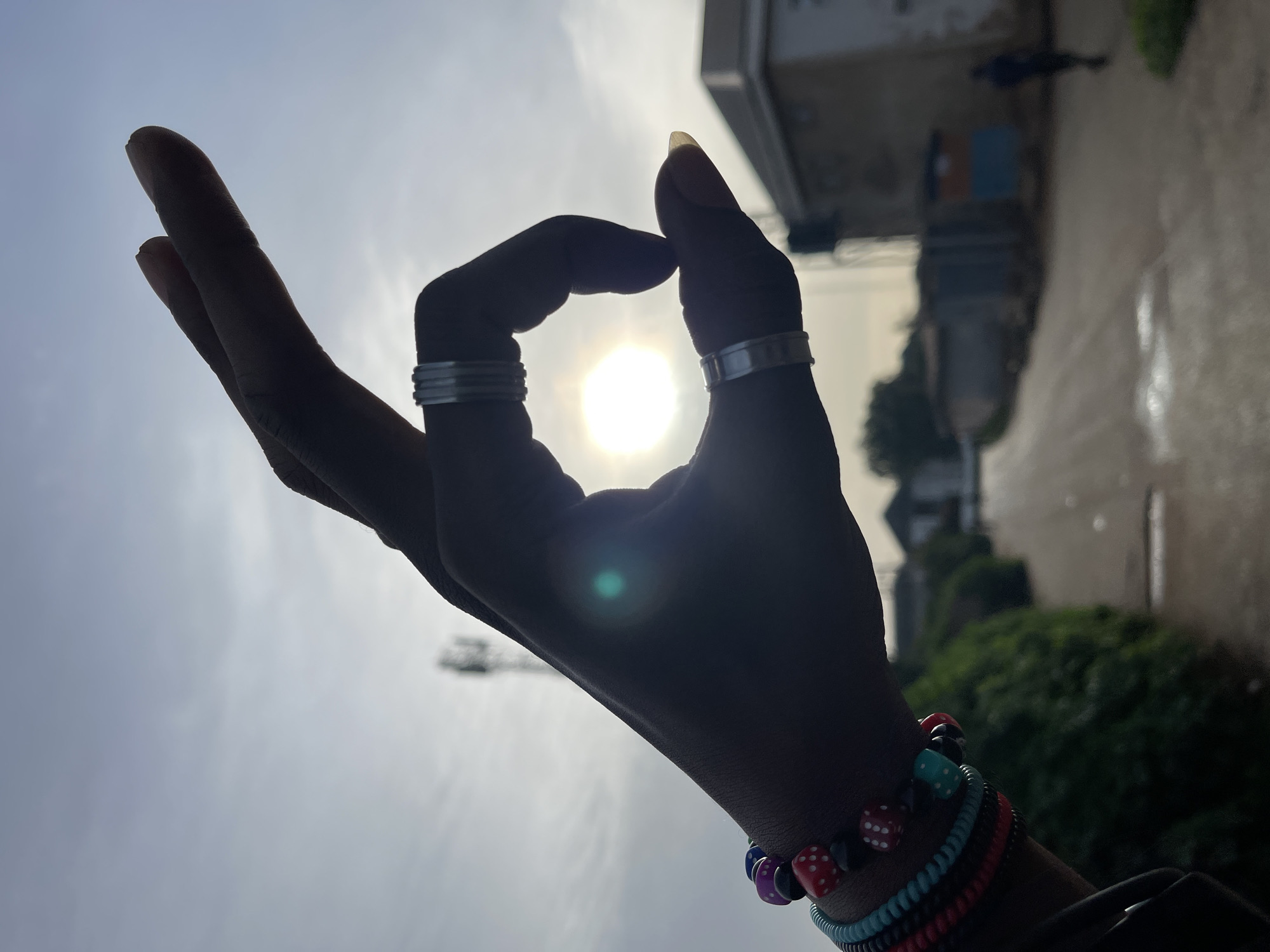 The image is a close-up, silhouette shot of a person's hand forming an "O" shape against a bright, hazy sky. The hand is adorned with several colorful beaded bracelets, including some with dice-shaped beads, and two silver rings on the fingers. The sun is visible behind the hand, creating a lens flare. In the background, a road stretches into the distance, flanked by green bushes and some low-rise buildings. A distant cell tower is also visible against the sky. The overall scene suggests an outdoor setting in what appears to be a residential area, possibly in Jos, Nigeria, given the context. The lighting and the presence of the cell tower are consistent with an urban or semi-urban environment in that region.
