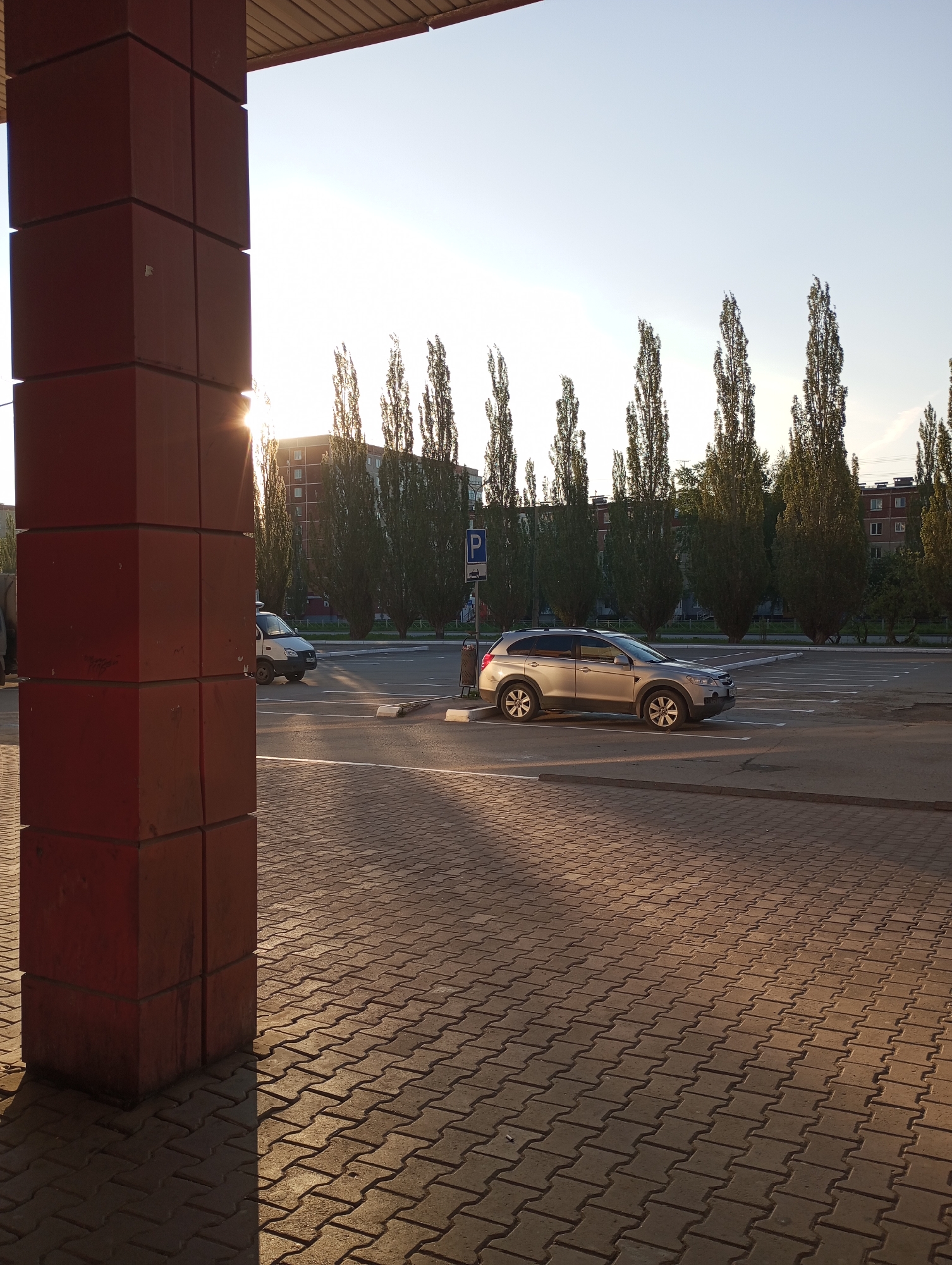 The photo shows a parking lot in Kamensk-Uralsky, Russia. A tan SUV is parked in one of the spaces. Behind it, a row of tall, slender trees lines the perimeter of the parking area. In the background, a multi-story brick building is visible. The foreground features interlocking paving stones, and a large, red, pillar-like structure stands to the left of the frame, partially obscuring a white van in the distance. The sun is low in the sky, casting long shadows across the parking lot. A parking sign is visible behind the SUV. The overall setting suggests an urban environment in the late afternoon or early morning.