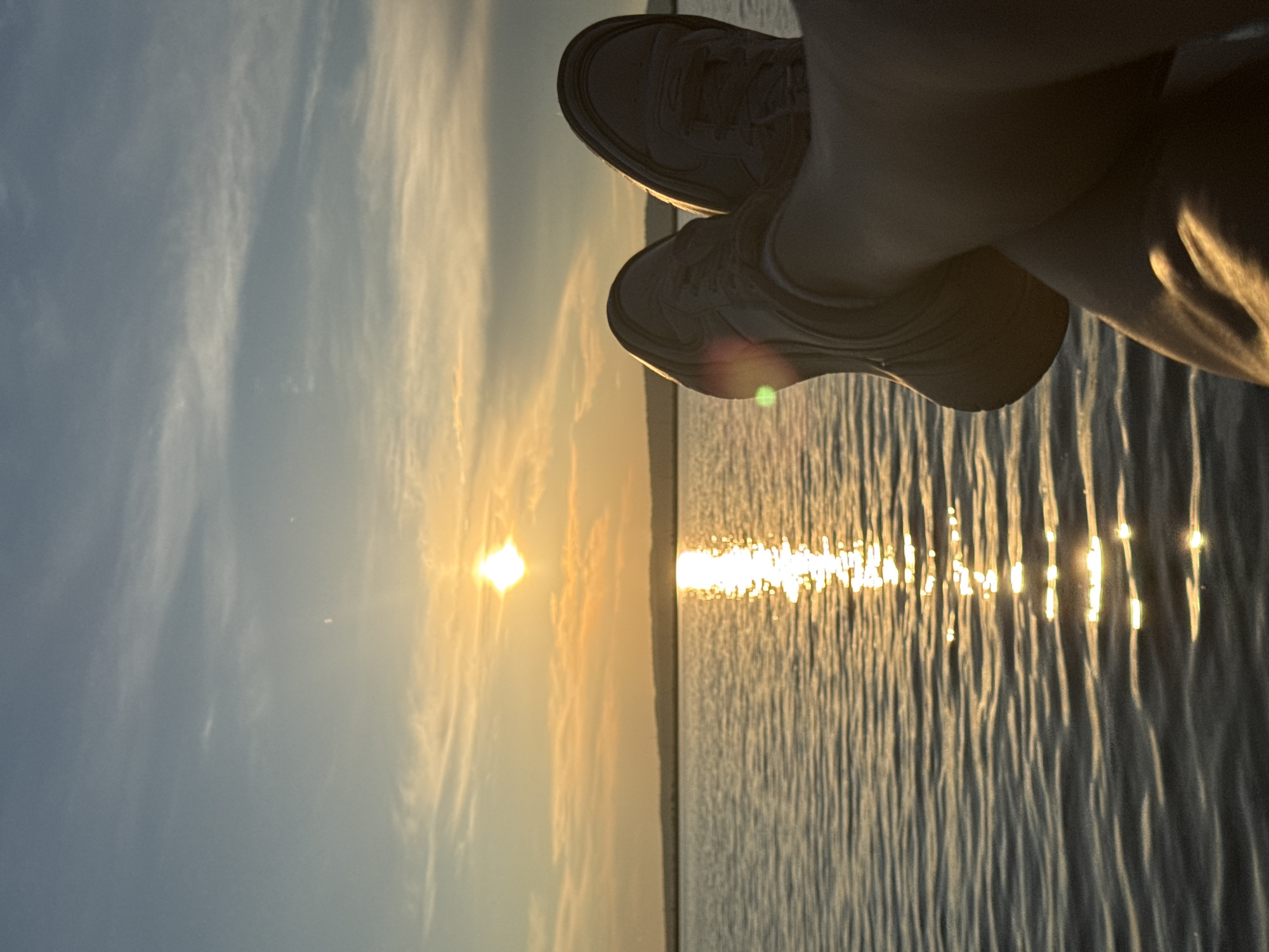 The image shows a close-up of a person's legs and feet, wearing light-colored sneakers, resting in front of a sunset over a body of water. The sun is low on the horizon, casting a warm golden light and a reflection on the rippling water. The sky is a mix of blue and orange hues with wispy clouds. The distant shoreline is visible as a dark, silhouetted line against the sky. The overall impression is one of relaxation and enjoying the natural beauty of the sunset. The location is the Kama River near Chaikovsky, Russia.