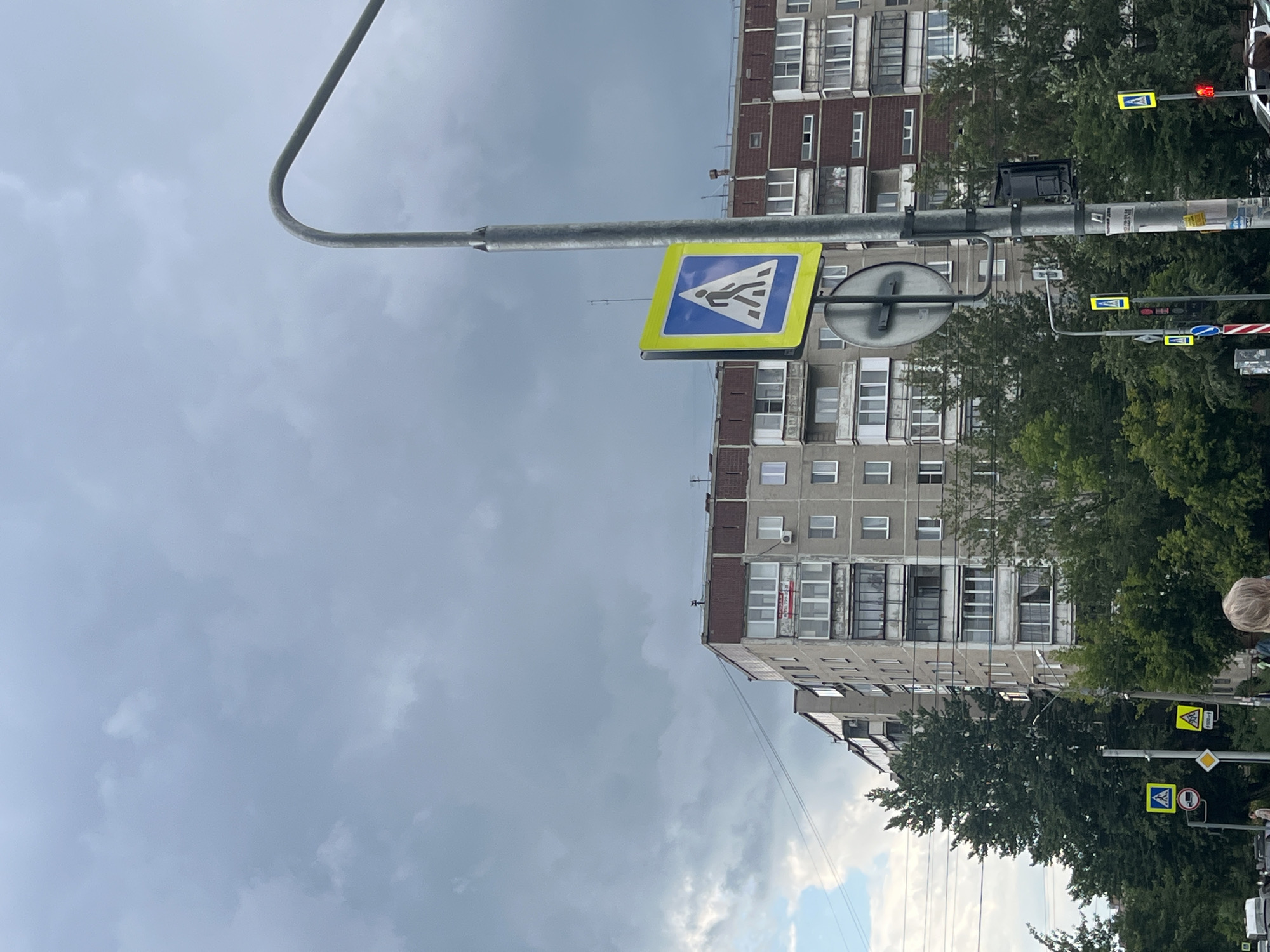 This media file depicts an outdoor scene in Chelyabinsk, Russia, under a cloudy sky, suggesting it might be a slightly overcast or stormy day. The perspective is from a low angle, looking upwards towards several traffic lights and pedestrian crossing signs mounted on a metal pole.
Dominating the background are multi-story apartment buildings, characteristic of Soviet-era architecture, with rows of windows and balconies. Dense green trees line the front of the buildings, partially obscuring them and suggesting a residential area with established greenery.
Several pedestrian crossing signs, distinguished by their yellow triangular frames with blue backgrounds and white pedestrian icons, are prominently visible. One sign is particularly clear, showing a stylized figure walking across a crosswalk. Traffic lights, some showing red and others unlit, are also mounted on the same pole system, indicating an active intersection. A circular traffic sign with a red border and a diagonal line is also visible, possibly indicating a prohibition.
A portion of a person's head and shoulder, with blonde hair, is visible at the far right edge of the frame, suggesting someone is present in the scene, likely observing or interacting with the environment. The overall impression is of an urban streetscape on a cloudy day, focusing on traffic control and pedestrian infrastructure within a residential setting. The lighting is diffused due to the cloud cover, and there are no strong shadows. Some stickers or labels are affixed to the traffic pole, but their text is not legible.