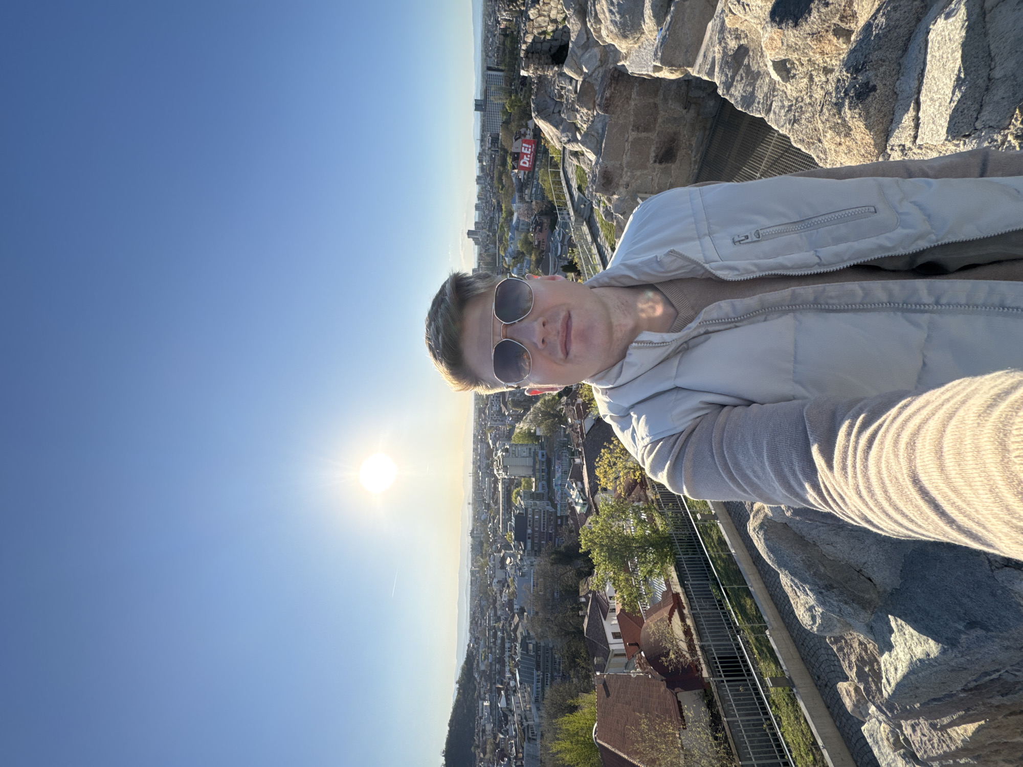 The image is a selfie taken from a high vantage point in Plovdiv, Bulgaria. A man wearing sunglasses and a light-colored jacket is the main subject. He appears to be standing on a stone structure, possibly a part of Nebet Tepe, overlooking the city. Below, a cityscape is visible, showing buildings, trees, and possibly houses. The sun is shining brightly, creating a glare in the sky. A "Dr.E!" billboard is visible in the city. The sky is clear and blue.