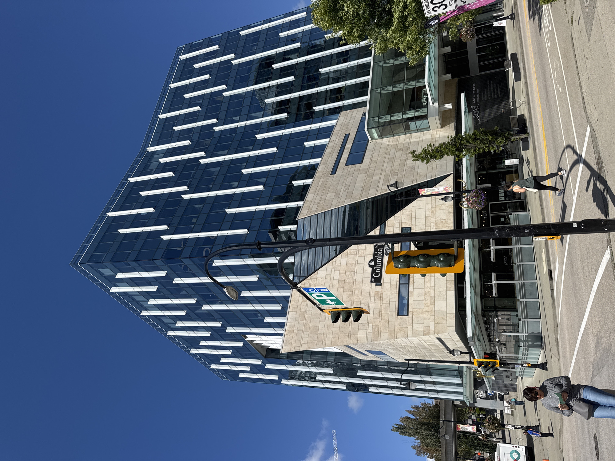 The image shows a street intersection in New Westminster, Canada, on a sunny day. A modern glass-fronted building with white vertical accents stands prominently on the right side of the frame. In the foreground, a woman in a grey sweater and blue jeans is looking down at a piece of paper while standing at a crosswalk. A traffic light is visible, and a street sign indicates "Columbia St". Further down the street, people are walking and a bus is parked on the left. The environment is urban, with clear blue skies and some trees.