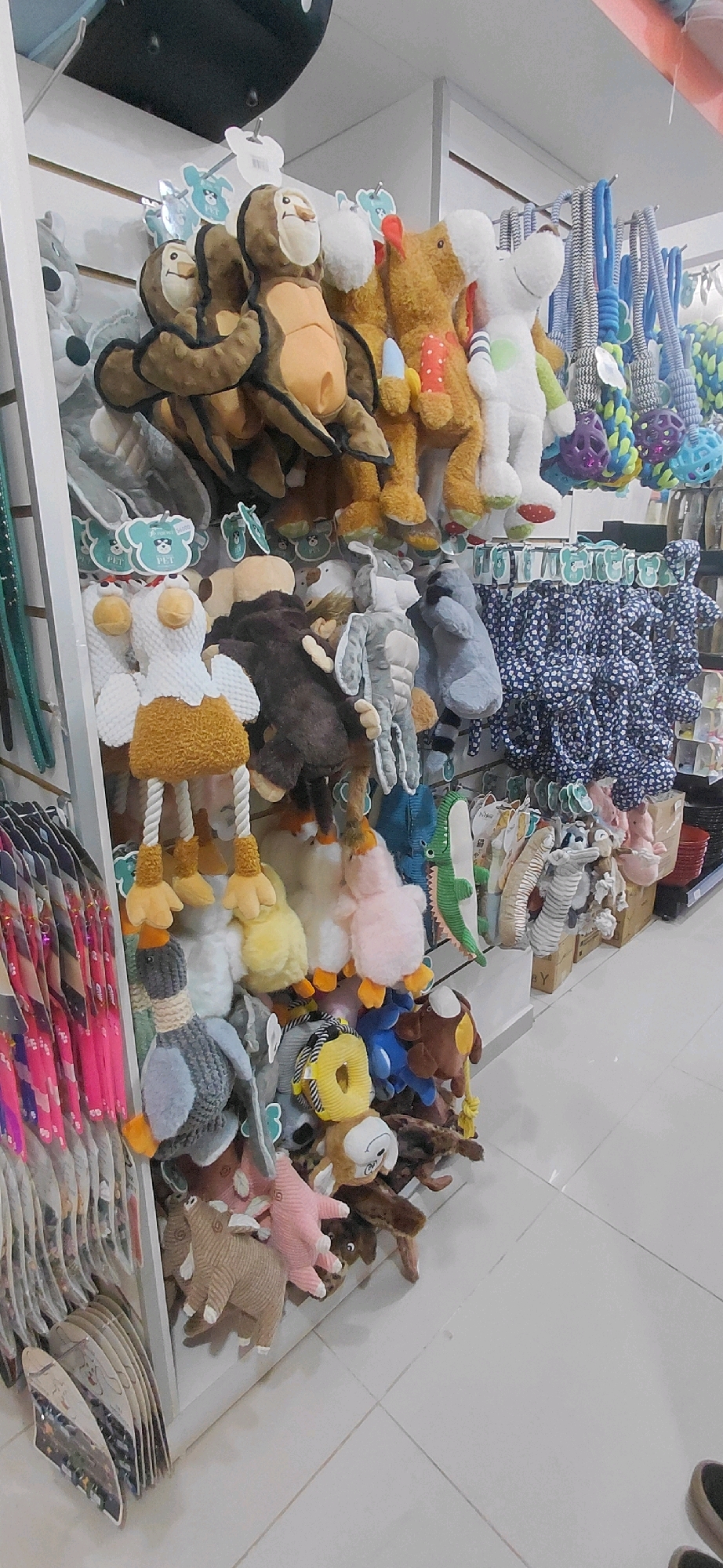 The image depicts a retail store display of assorted plush animal toys and dog leashes in Encarnación, Paraguay. The merchandise is arranged on white shelving units and hooks, with some items hanging freely. Prominently featured are several stuffed monkeys, a brown bear, a white dog, a yellow duck, a pink duck, a gray duck-like toy, and a green crocodile toy. Various dog leashes and collars, made of braided material and colorful fabric, are also visible. The background includes white walls with horizontal shelving lines, and the floor is tiled in a light color. The overall lighting is bright, suggesting an indoor commercial setting. No people are visible in the image.