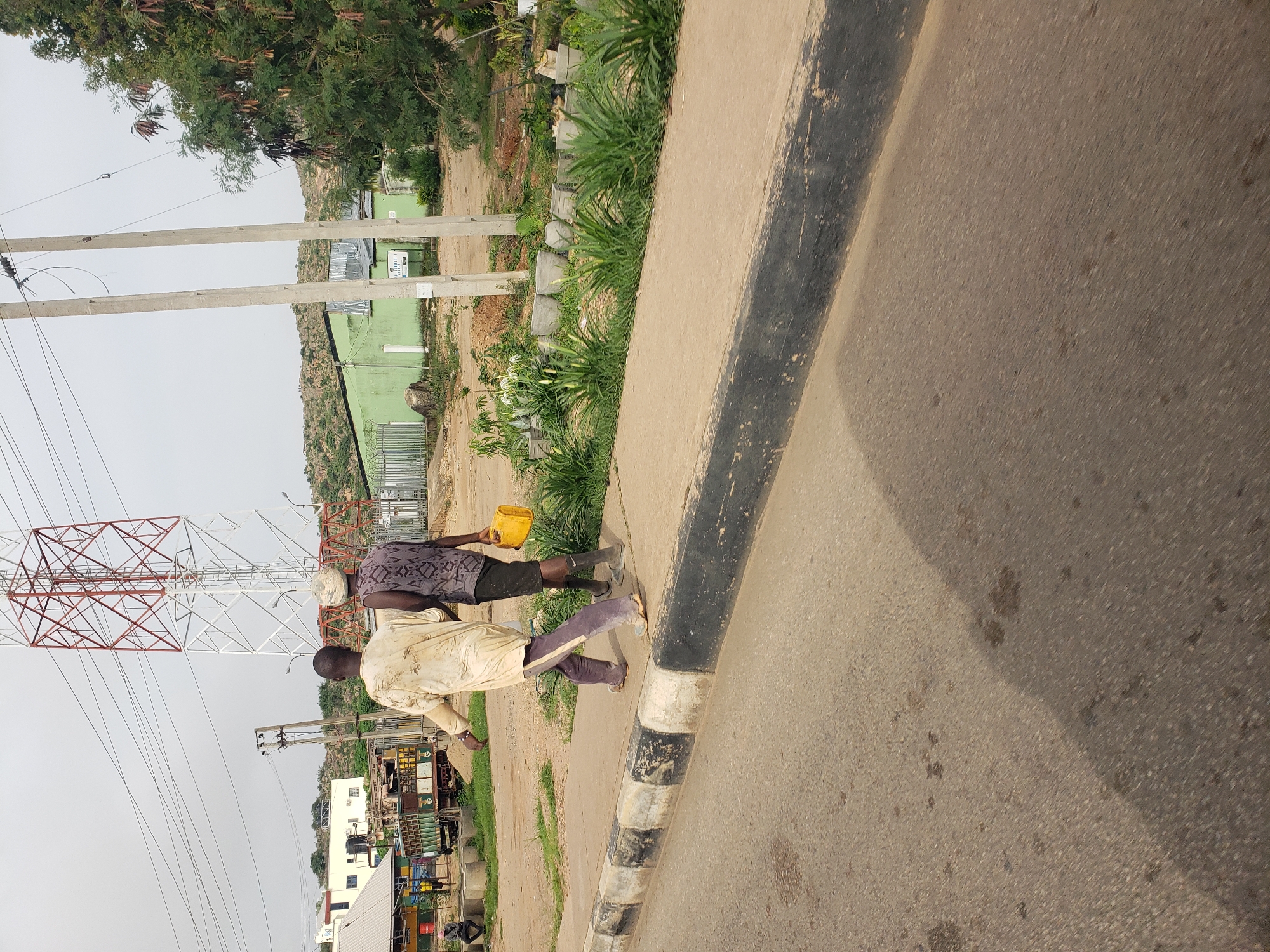 Two men are walking on a sidewalk next to a road in an outdoor urban setting. The man on the left is wearing a light-colored, loose-fitting garment and dark pants, and is walking barefoot. The man on the right is wearing a dark patterned sleeveless shirt, dark shorts, and boots, and is carrying a yellow plastic container. Behind them, a large red and white transmission tower is visible, along with power lines, a green building, a truck, and a fence. The ground is paved with asphalt, and the sidewalk has a black and white striped curb. The sky is overcast.