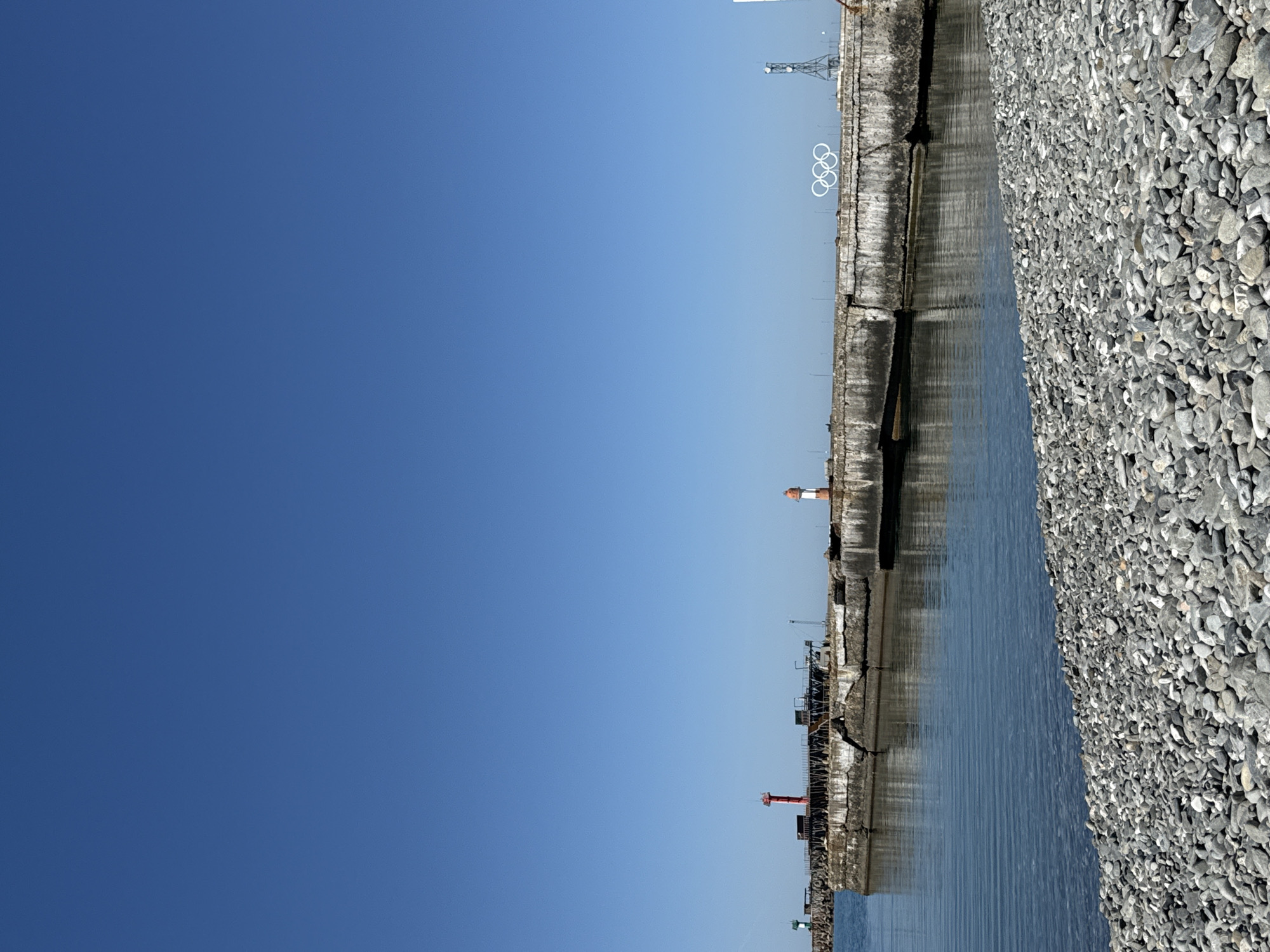 The image, when viewed upright, captures a serene coastal or harbor scene under a vast, cloudless blue sky, which dominates the upper half of the frame. Running horizontally across the mid-ground is a concrete pier or breakwater, separating the sky and some structures from the calm, blue water below. To the right of the water, a wide stretch of grey, rounded pebbles forms a bank or beach, filling the right side of the image.
Along the concrete pier, several distinct features are visible. A prominent white sculpture of the five interlocking Olympic rings stands out against the sky in the upper right quadrant. Further along the pier, three small lighthouses or beacons can be identified: one with a white and orange top, one with a red top, and another with a green top. A tall, metal lattice tower, resembling a communication tower or a crane structure, is partially visible in the upper right background. Additional smaller, dark industrial-like structures are scattered along the pier's length.
No people or discernible activity are present, giving the scene a tranquil atmosphere. The bright, clear conditions indicate it is daytime, likely late morning or early afternoon. The presence of the Olympic rings strongly suggests a location associated with the Olympic Games. No legible text is visible within the image.