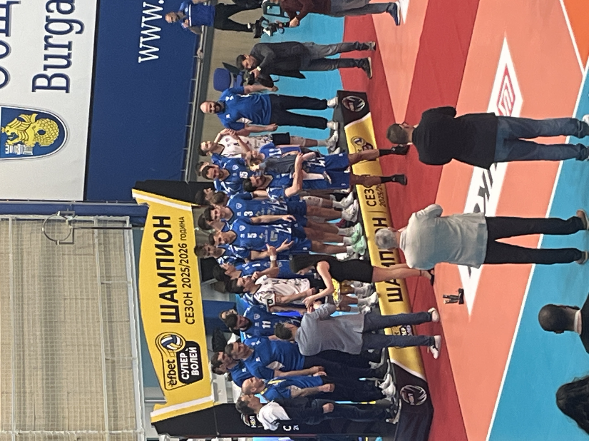 The image shows a group of people, mostly men, posing for a photo. They appear to be a sports team, wearing blue and white jerseys with numbers. A yellow banner stands to their left, displaying the text "ШАМПИОН СЕЗОН 2025/2026 ГОДИНА efbet СУПЕР ВОЛЕЙ." There are additional people around the team, presumably taking photos or observing. The background includes what appears to be a banner displaying "Burga" and "www." The image is taken at floor level, showing the red and blue pattern of the volleyball stadium flooring.