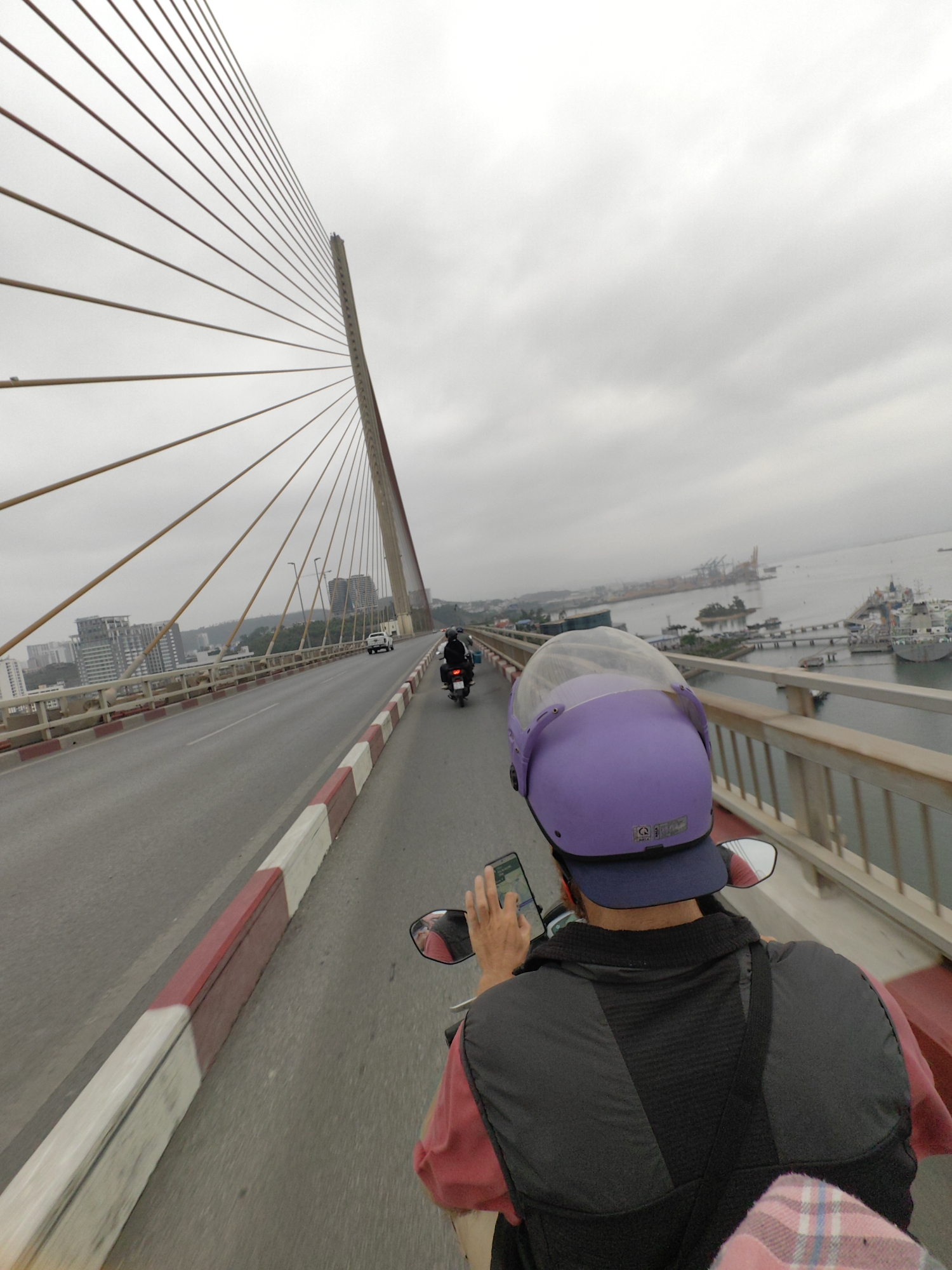 The image shows a person riding a motorcycle across a bridge in Hong Gai Ward, Vietnam, as indicated by the cityscape and port facilities visible in the background. The person is wearing a purple helmet and a dark vest over a pink shirt. The person is holding a phone, potentially using it for navigation. The bridge is a cable-stayed bridge with a prominent tower and numerous cables extending to the bridge deck. Other vehicles, including a motorcycle and a car, are visible further ahead on the bridge. The weather appears overcast.