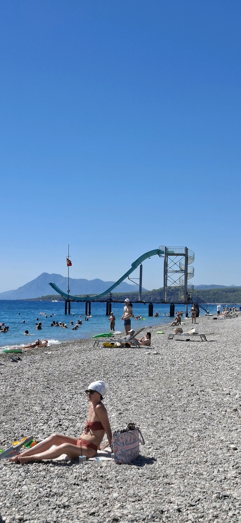 A wide-angle, vertical shot captures a sunny day at a pebble beach in Kemer, Turkey. The clear blue sky dominates the upper portion of the frame. In the middle ground, a turquoise water slides down from a tall metal structure. Numerous people are visible, swimming and lounging in the sea and on the beach. A Turkish flag flies from a pole near the water slide. In the foreground, a woman in a red bikini and white headscarf sits on a towel on the pebble beach, looking to her left. Adjacent to her is a patterned beach bag. The overall scene depicts a lively and recreational beach environment.