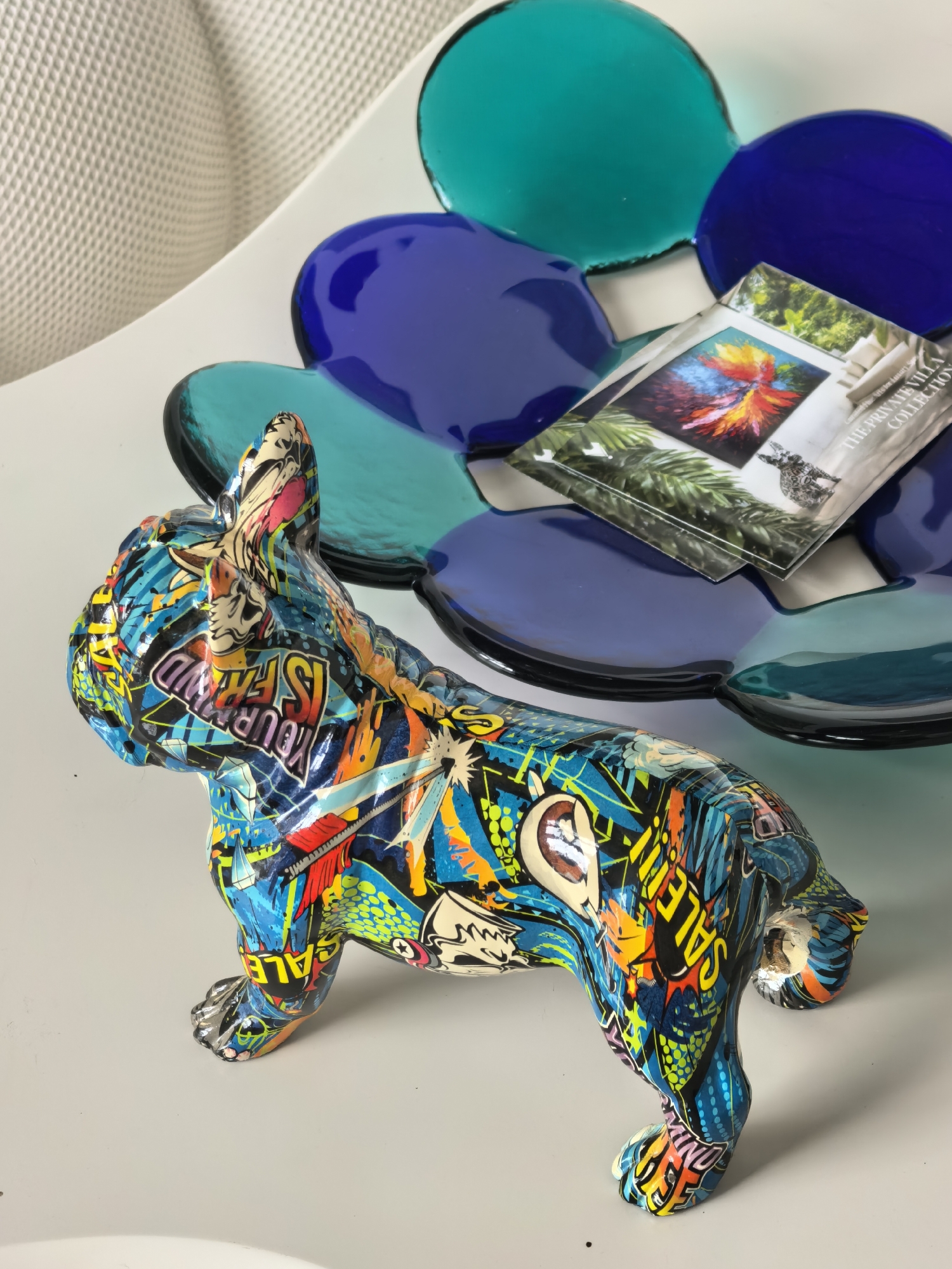 A vibrantly decorated French bulldog figurine, covered in a collage of graffiti-style art featuring bold colors and text, is positioned on a white surface. Behind the figurine, a cluster of translucent, multi-colored glass plates, arranged in a flower-like pattern, creates a backdrop. The plates range in color from deep blue to teal and purple. A small booklet or card with images of artwork and text is resting on the glass plates. The overall scene suggests an indoor setting, possibly a home or a shop display, with a white textured material visible in the upper left corner, implying a wall or fabric. There are no people or discernible actions taking place. The location in Ko Samui District, Thailand, is not evident from the visual information provided in the image.