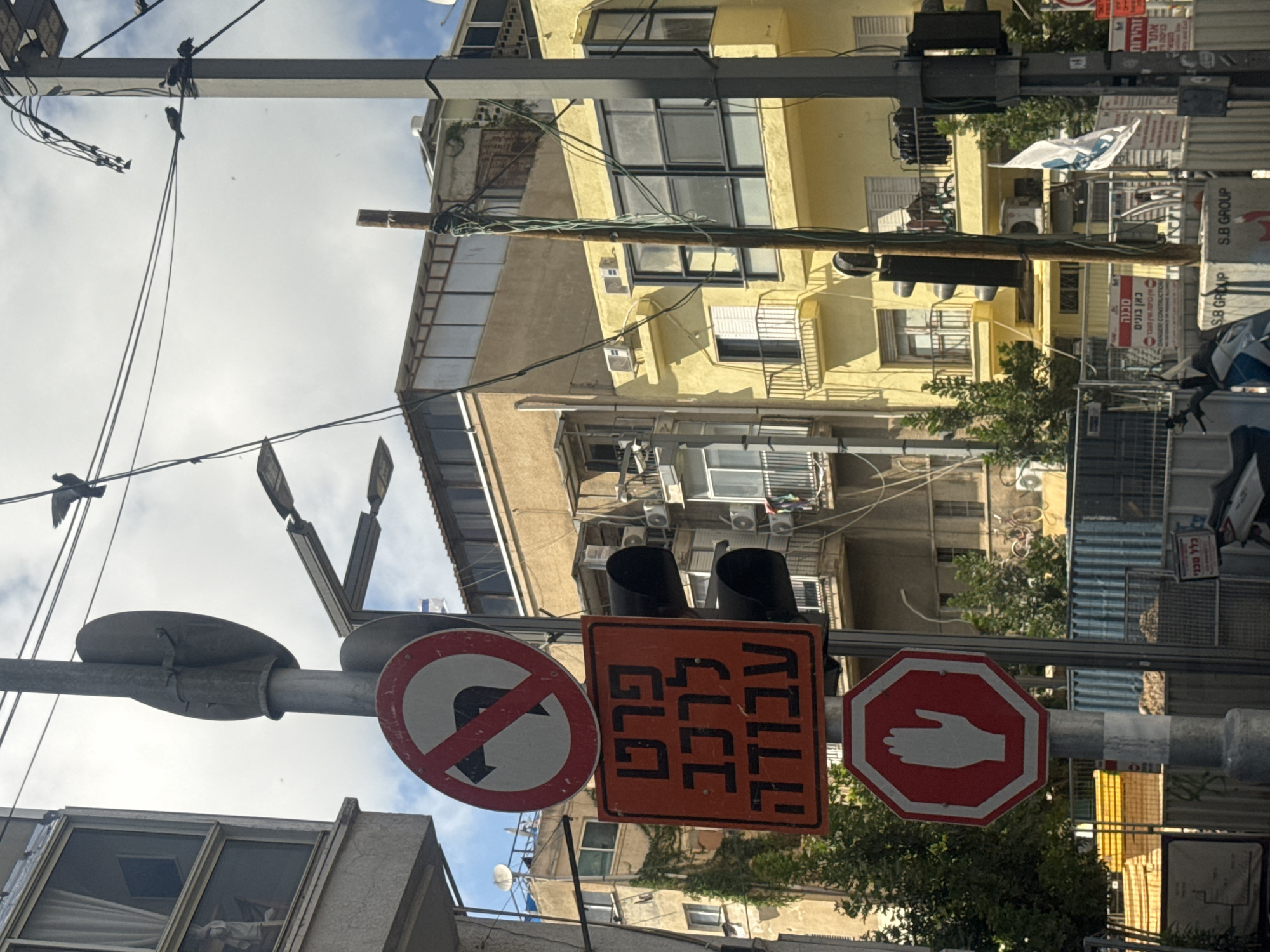This eye-level photograph is a street-level scene in Tel-Aviv, Israel, depicting an array of signs mounted on a metal pole. The signs include a red and white "no U-turn" sign, an orange rectangular sign with Hebrew writing indicating "work ahead," and a red and white octagonal sign with a white hand symbol to indicate "stop." The signs are in the foreground and slightly angled towards the camera. In the background are several buildings, some painted yellow, with numerous windows and air conditioning units visible. There are also overhead wires, street lights, and vegetation visible. The sky is partly cloudy. The scene has a busy, urban feel.