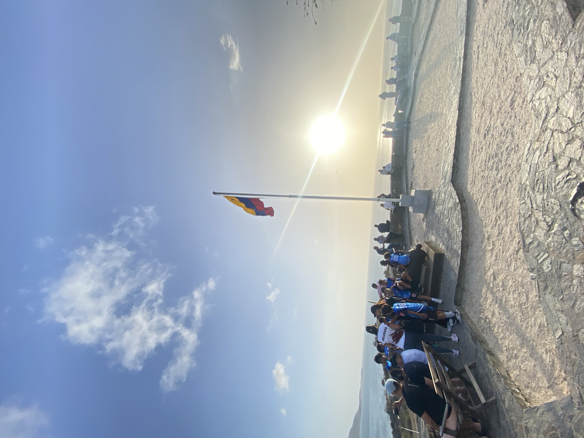 The image shows a horizontal outdoor scene on the oceanfront promenade in the city of Juan Griego, Venezuela. A large group of people are gathered on a stone-paved area, with some sitting and others standing. Many are wearing blue sports jerseys. A Venezuelan flag is waving atop a tall pole in the mid-ground. The sky is mostly clear blue, with a few clouds. The sun is shining brightly, creating a hazy, bright area in the sky. The horizon line shows the ocean meeting the sky. A low stone wall is visible.