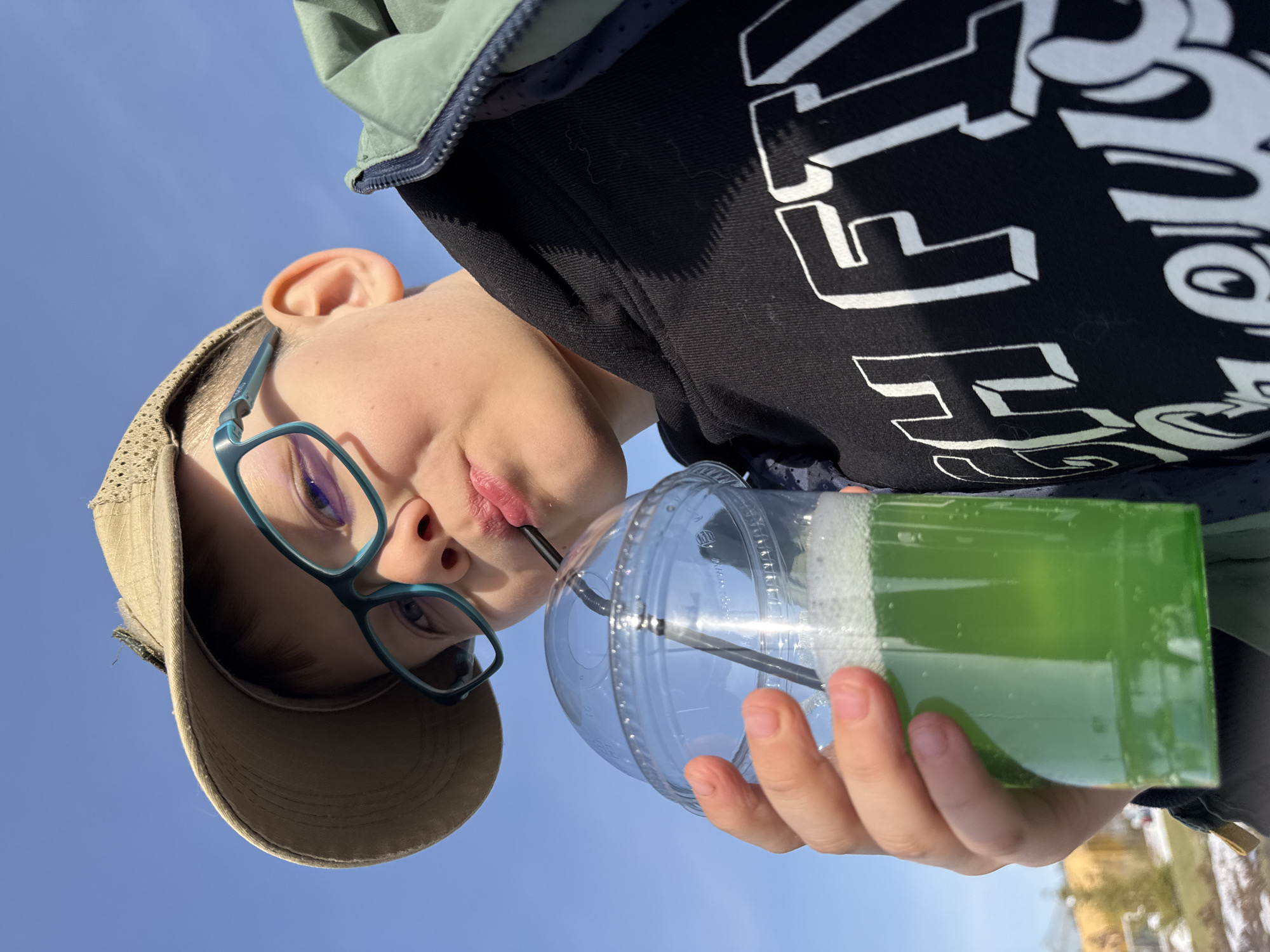 A low-angle medium shot shows a fair-skinned boy in Perm, Russia drinking a green beverage with a straw from a clear plastic cup. He wears blue glasses and a tan baseball cap. He is wearing a black hoodie with white writing. He wears a green jacket. The background is a clear blue sky. A building is visible at the bottom edge of the image.