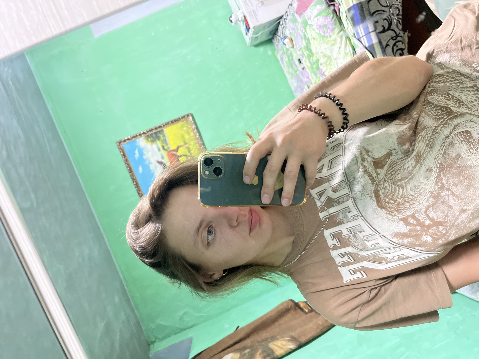 A young woman with light brown hair is taking a mirror selfie in a room. She is wearing a light brown t-shirt with a graphic print and a silver chain necklace. She has two spiral bracelets on her left wrist. The room has light green walls, and a framed picture of a horse in a field hangs on one wall. To the right of the woman, a bed with patterned bedding is visible. The image appears to have been taken indoors, possibly in a residential setting. No specific location within Nizhny Tagil, Russia can be determined from the visual information provided.