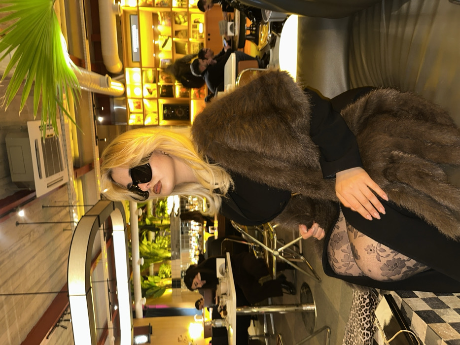 A fair-skinned blonde woman wearing black sunglasses and a fur wrap is seated indoors. She is wearing patterned tights and a black dress. The background includes a bar with patrons, plants, and lighting fixtures. The room has high ceilings and industrial elements like ducts. A leafy green plant is visible in the upper left corner. The image appears to be taken in a restaurant or lounge setting.