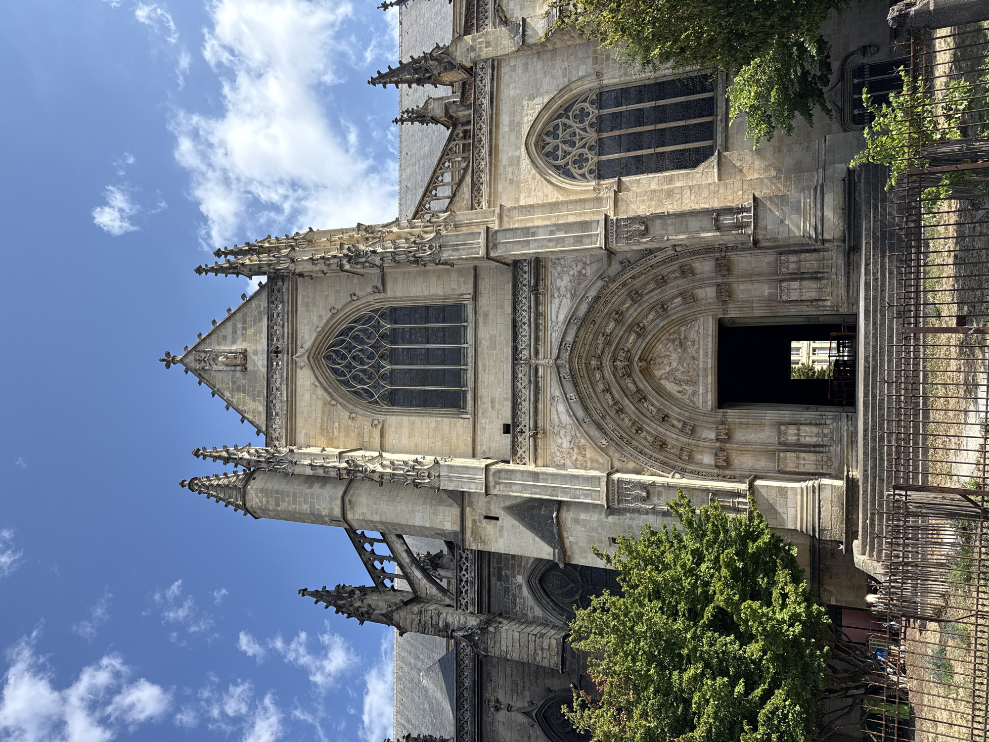 The image shows the exterior of a grand Gothic cathedral in Bordeaux, France. The cathedral is constructed of stone and features intricate architectural details such as pointed arches, gargoyles, and ornate carvings around the entrance. A large, circular stained-glass window is visible above the entrance.
The perspective is from a low angle, looking up at the cathedral against a bright blue sky dotted with fluffy white clouds. Lush green trees frame the right side of the building, with branches extending towards the stone facade. A rustic metal fence runs along the right edge of the image, partially obscuring a set of stone steps leading to the cathedral's entrance. Through the open doorway, glimpses of the interior can be seen, including more architectural elements and possibly some lighting fixtures.
The lighting suggests it is daytime, with the sun casting shadows on the building's facade. The overall mood is one of awe and admiration for the historical architecture. There is no visible text in the image.