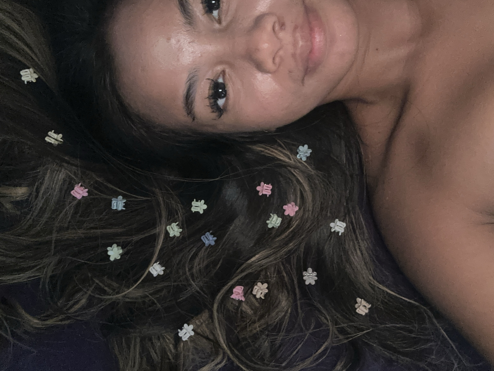 A woman with long, wavy brown hair with blonde highlights lies on a dark blue surface. Scattered throughout her hair are small, colorful flower and bow-shaped hair clips. The woman faces the camera with a slightly tilted head. The lighting is soft, illuminating her face and hair. The background is not discernible. There is no evidence to suggest this image was taken in North Jakarta, Indonesia.