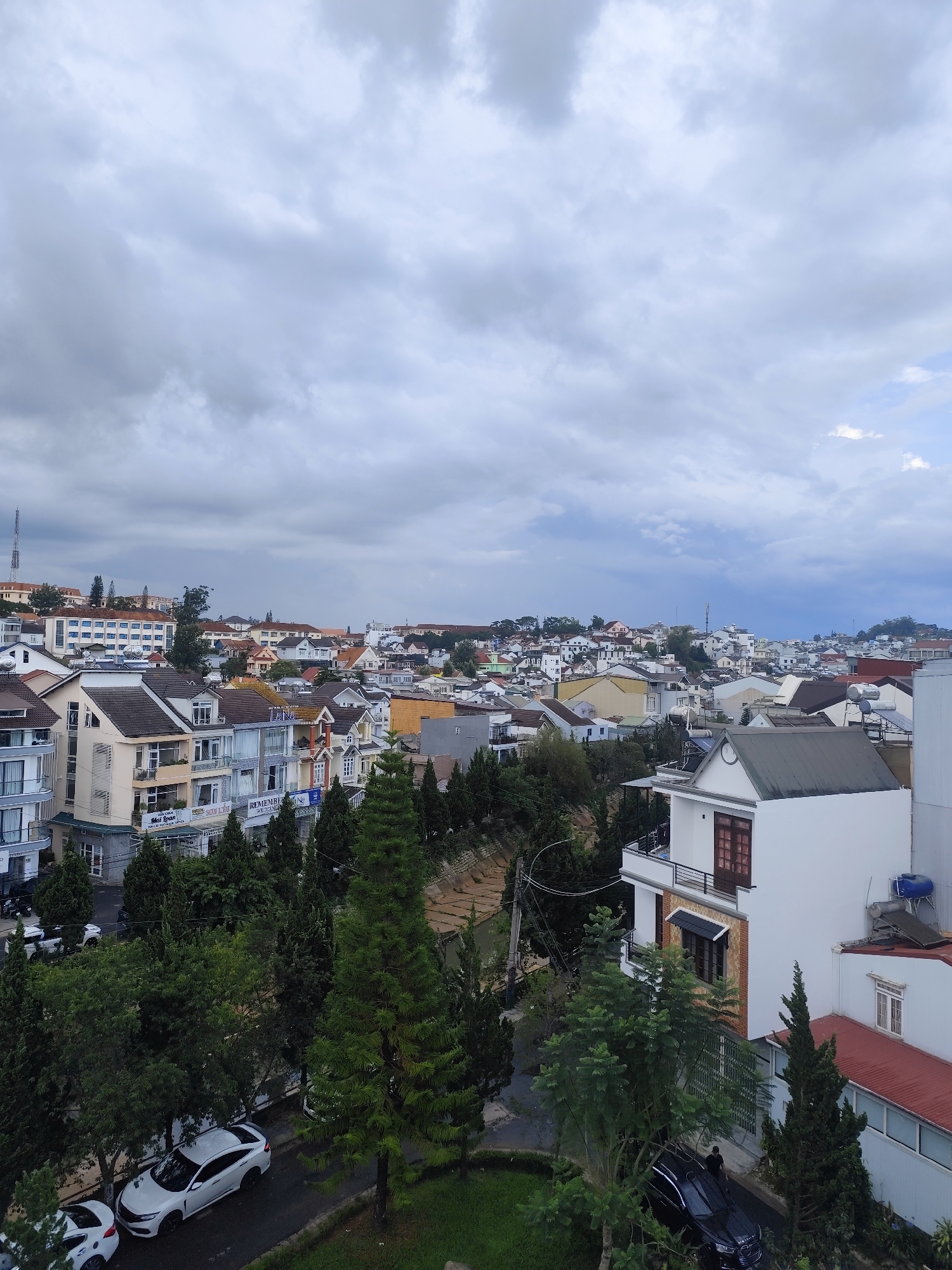 The image captures a cityscape in Da Lat, Vietnam, under an overcast sky. The scene is dominated by a mix of residential buildings, ranging from multi-story structures to smaller houses with pitched roofs. The buildings feature a variety of architectural styles and colors, typical of the urban landscape in Da Lat.
In the foreground, dense greenery including trees, shrubs and grass partially obscures a street with parked cars. Among the cars are at least two white sedans. A black SUV sits in front of a multi-story building. There is no indication of people or actions directly visible.
The overall environment suggests a high-density residential area, with the dense building arrangement reaching the distant horizon where buildings fade into the background.