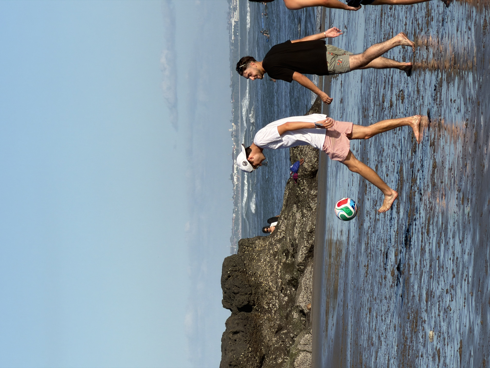 The image, taken in North Kuta, Indonesia, shows two young men on a rocky shore. One man, wearing a black t-shirt and patterned shorts, stands with his head bowed and a slight smile on his face, appearing to be looking down at the wet sand. He has sunglasses perched on his head. The second man, in a white t-shirt and pink shorts, is bent over, reaching towards the ground with his left hand. He is wearing a white baseball cap. Both men are barefoot.
A multi-colored soccer ball is visible on the wet surface near the second man's outstretched hand. The wet ground, likely a beach or tidal area, reflects the sky and the surrounding environment. In the background, the ocean can be seen with waves and a distant coastline with buildings. The sky is a clear, bright blue, suggesting it is daytime, likely late morning or early afternoon, with good weather. The overall mood seems relaxed and casual, perhaps a group of friends enjoying their time by the sea. There is no visible text in the image.
